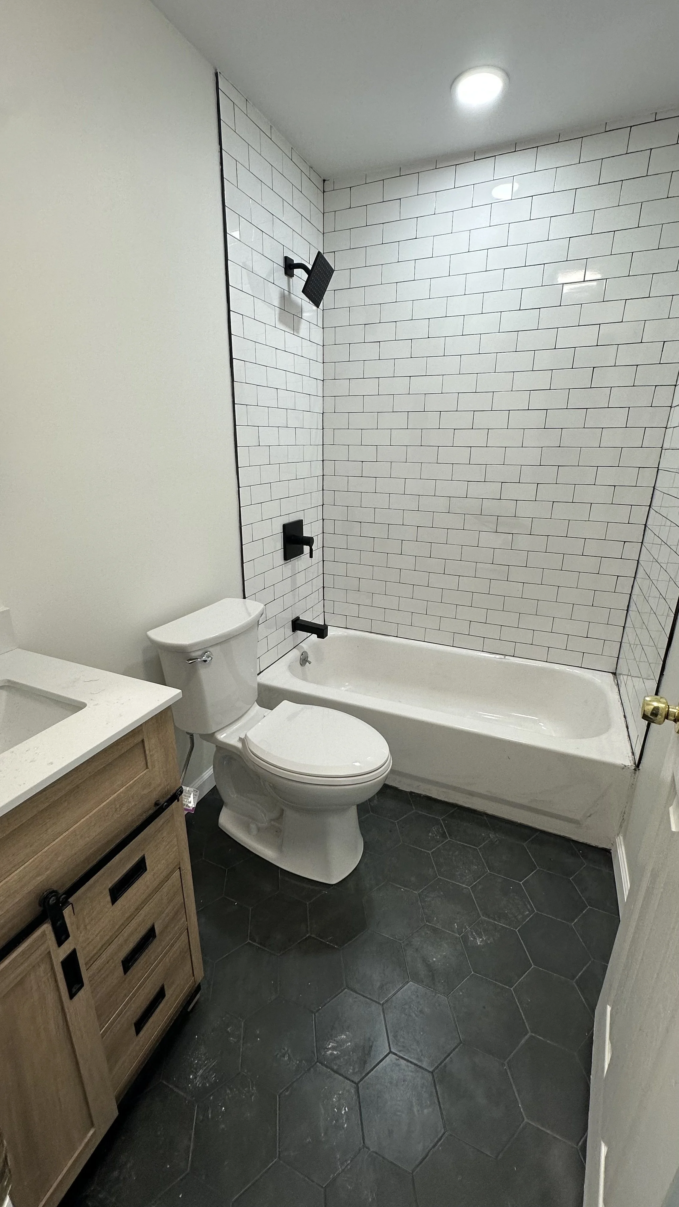 Bathroom with a toilet, a wooden vanity, and a bathtub with white subway tiles on the walls and gray hexagonal tiles on the floor.