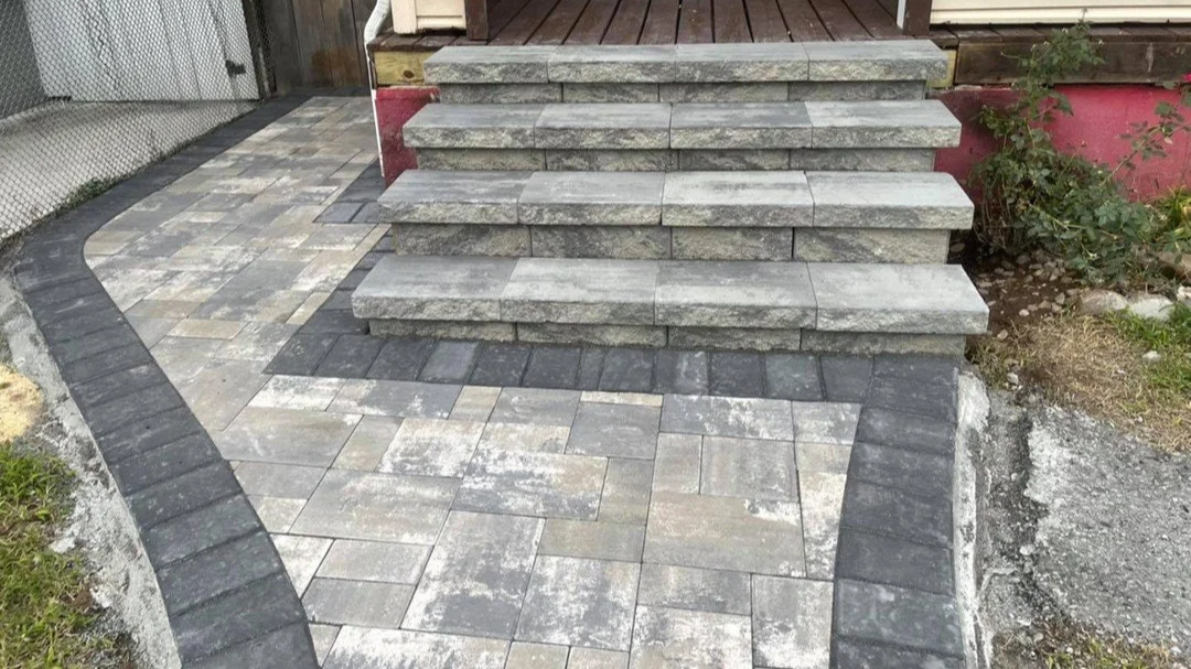Stone steps leading to a porch, with a newly paved walkway made of rectangular paving stones in gray and black, outlining a curved pattern.