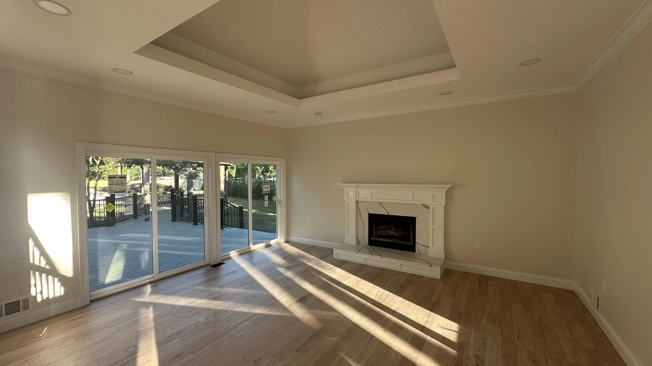 Emptyliving room with a fireplace, large sliding glass doors leading to an outdoor deck, hardwood floors, and sunlight casting shadows inside.
