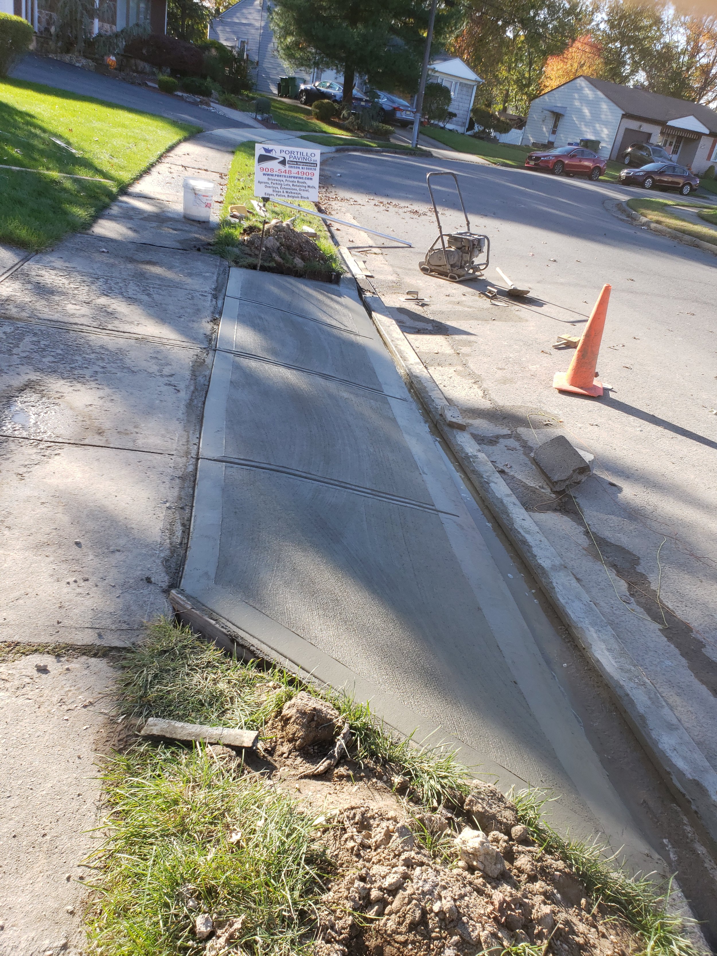 Sidewalk under construction with fresh concrete, construction tools, an orange safety cone, and a sign for paving services. Residential street in the background.