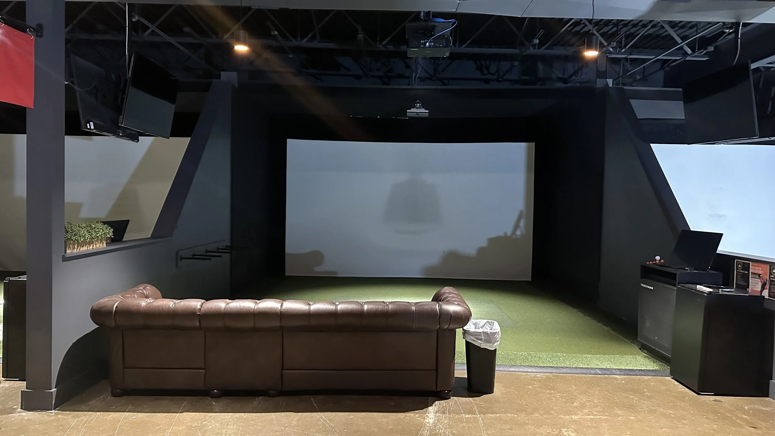 Indoor golf simulator room with large projection screen, leather sofa, and golf simulation equipment.