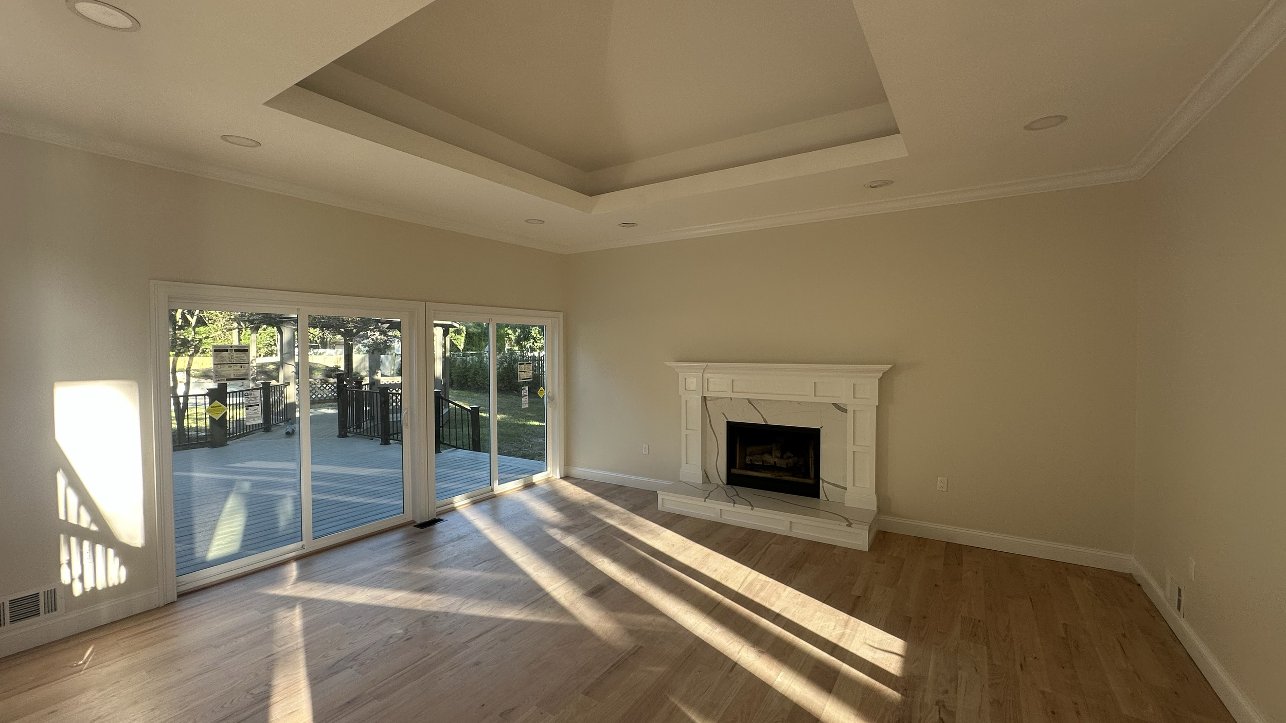 Empty living room with large sliding glass doors leading to a backyard patio, a white fireplace, and hardwood floors, illuminated by sunlight.