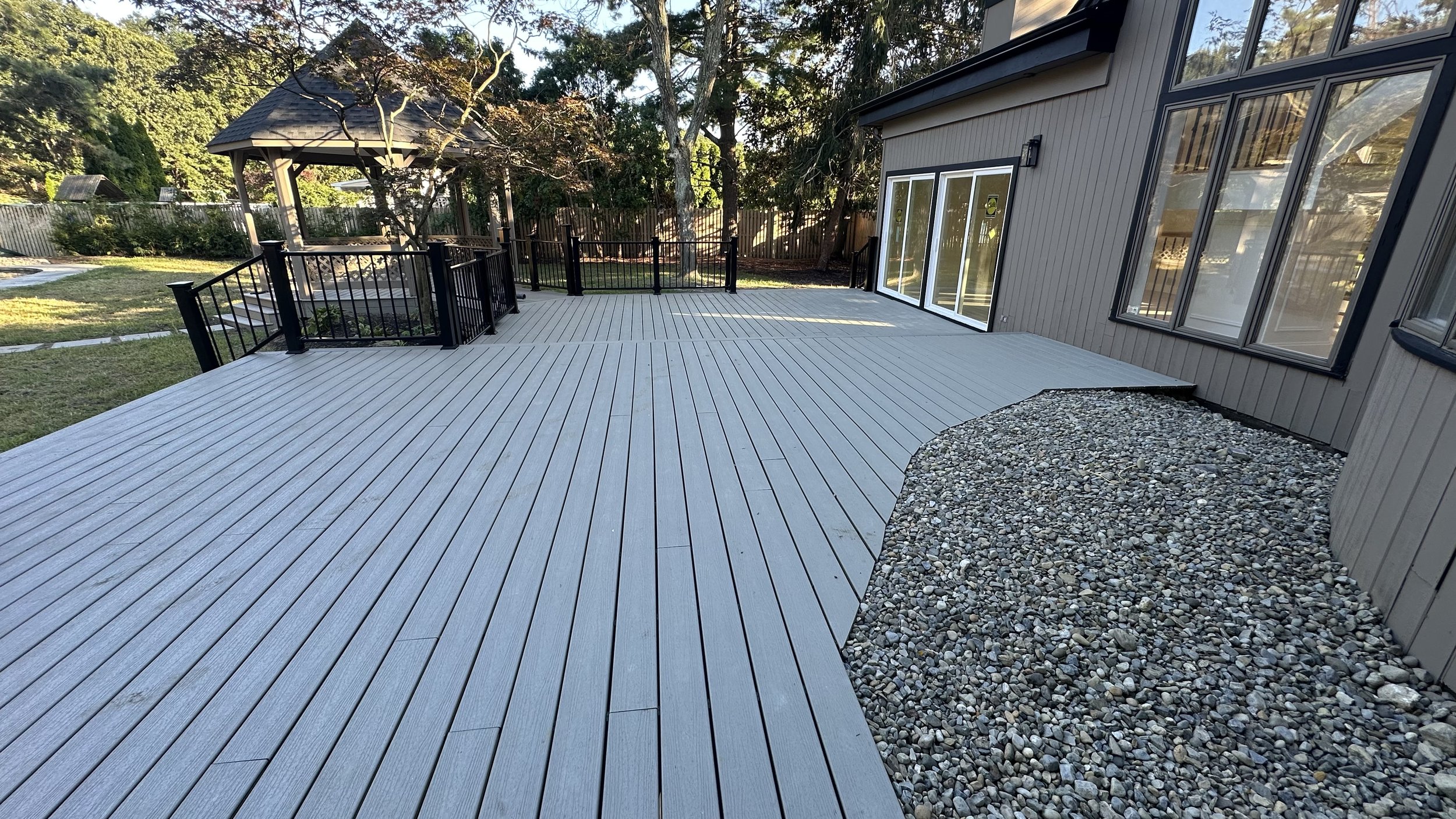 Photo of a backyard deck with gray wooden planks, a black railing, and a nearby gravel area. There is a house with large windows and sliding glass doors. The yard has trees and a gazebo in the background.