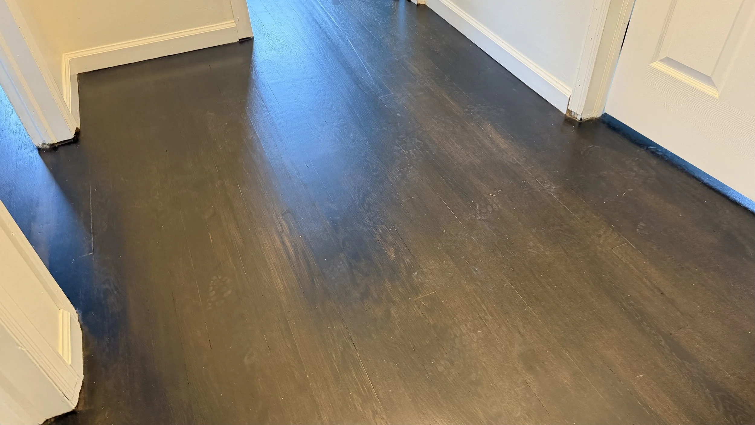 Close-up view of a dark wooden floor with water spots and streaks, next to a white door frame and beige wall.