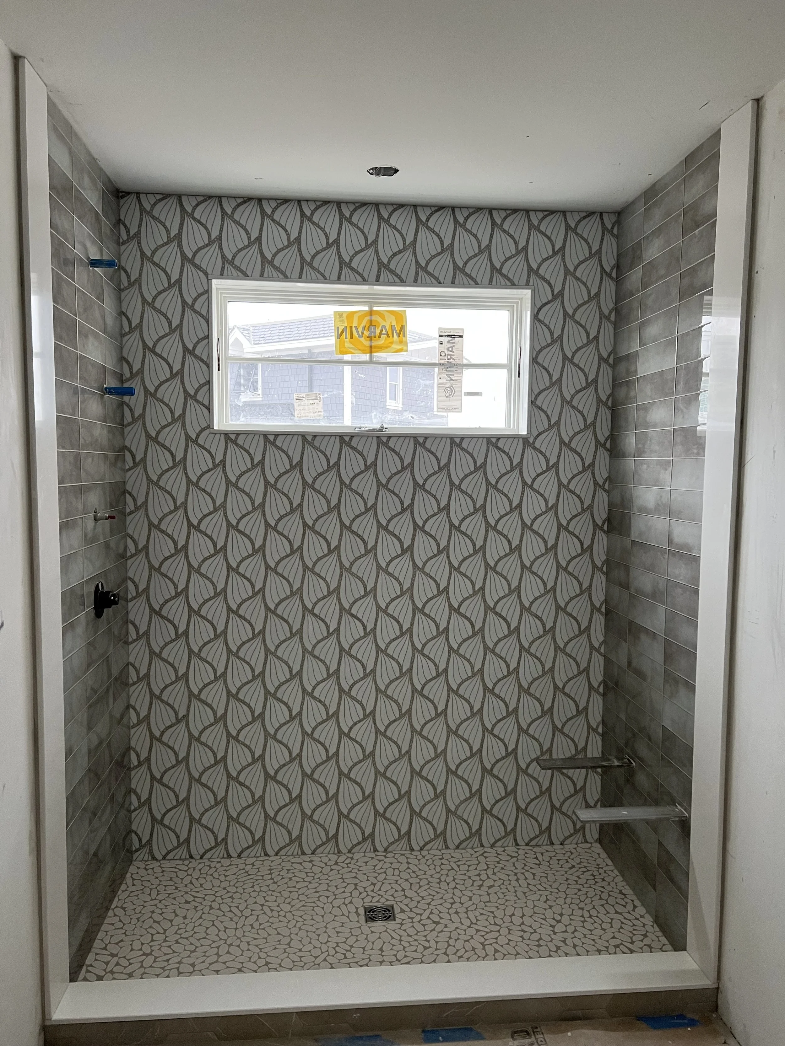 A shower area with patterned wall tiles, a window with a yellow warning sign, a drain on the floor, and unfinished corners.