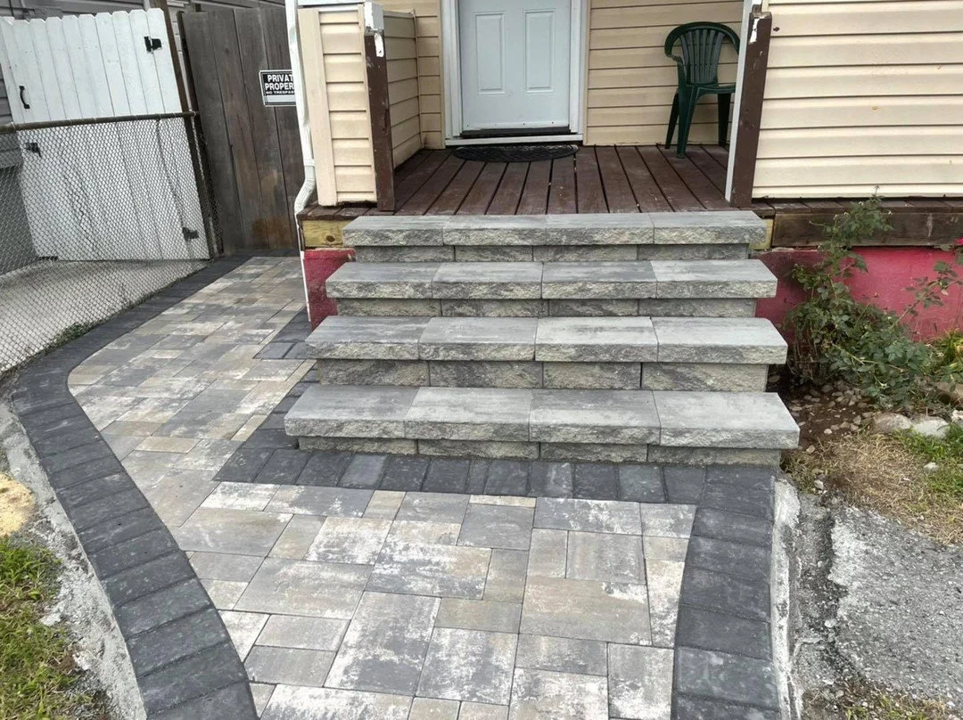 Stone steps leading to a small porch with a green chair, surrounded by a cleared yard with some plants and a paved walkway.