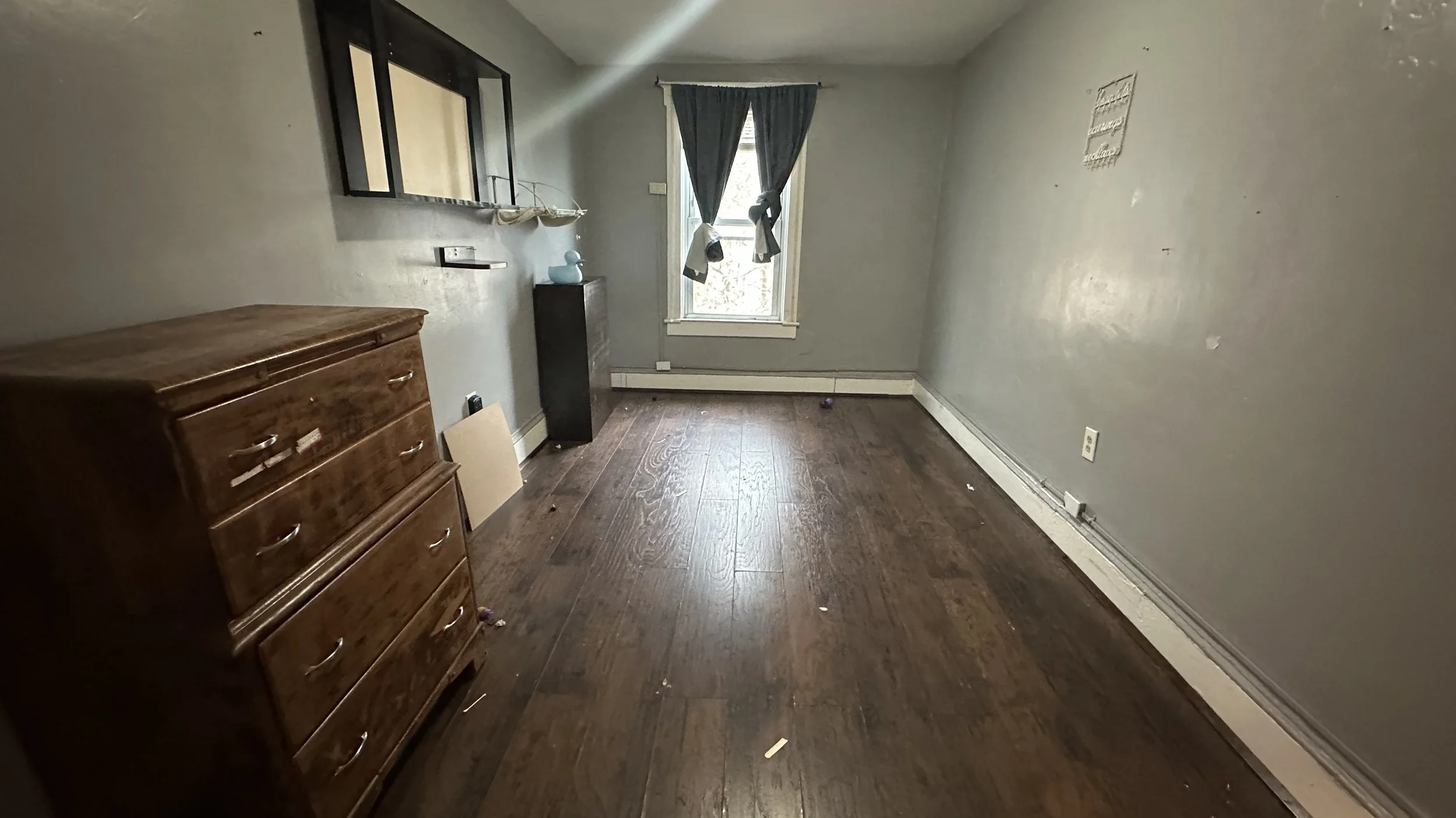 Empty room with a wooden dresser on the left, a window with curtains on the far wall, and some scattered debris on the dark hardwood floor.