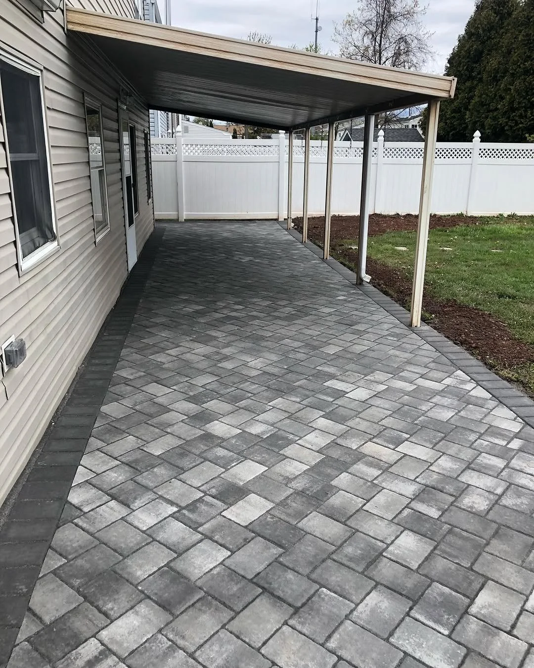 Backyard patio with interlocking gray and black stone pavers, covered by a metal roof, enclosed by a white privacy fence, next to a house with beige siding and windows.