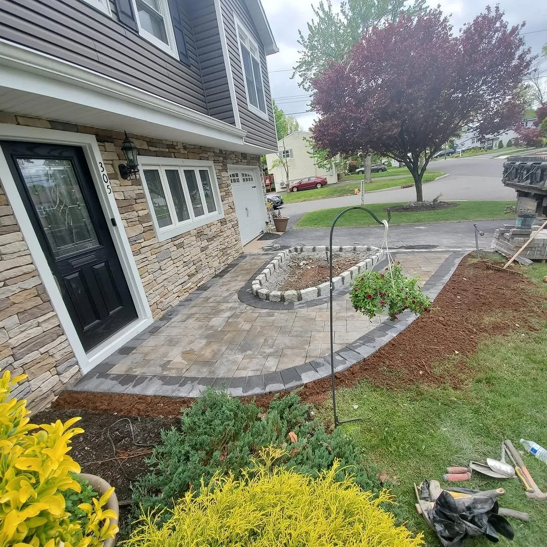 Front yard with a newly paved brick walkway, a flower bed with a hanging basket of flowers, a tree, and some construction tools.