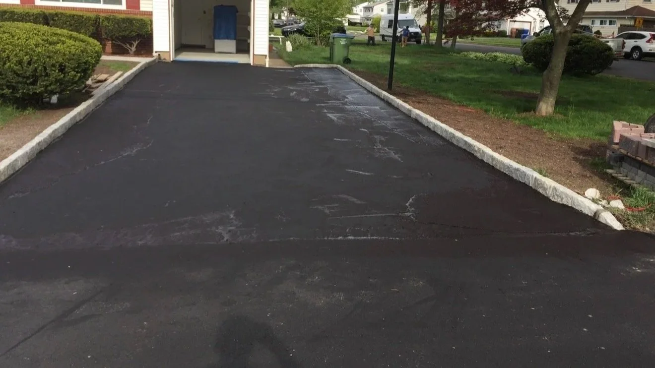 Freshly paved black asphalt driveway with white concrete edging in front of a house, surrounded by green grass, trees, and neighboring houses.