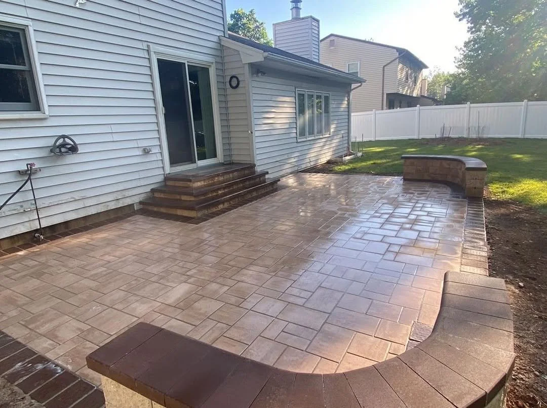 Newly installed stamped concrete patio with steps leading to sliding glass door of a house, surrounded by a white fence and neighboring houses, with some sunlight and greenery in the background.