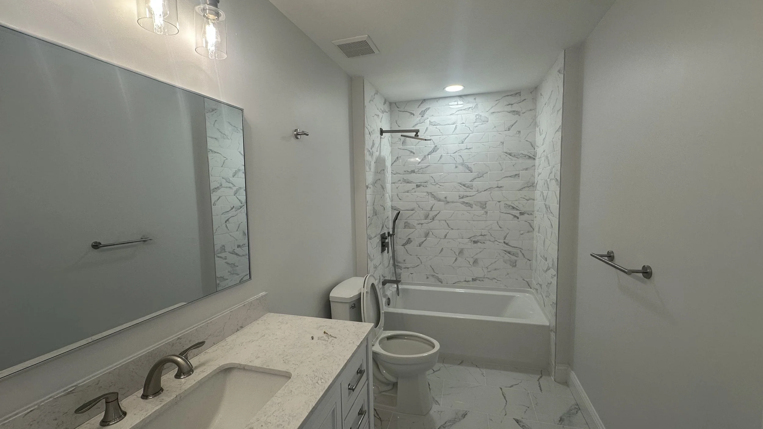 Modern bathroom with white marble tiles on the wall and floor, a bathtub with a showerhead, a white toilet, a white vanity with a sink and faucet, a large mirror, and towel racks on the walls.