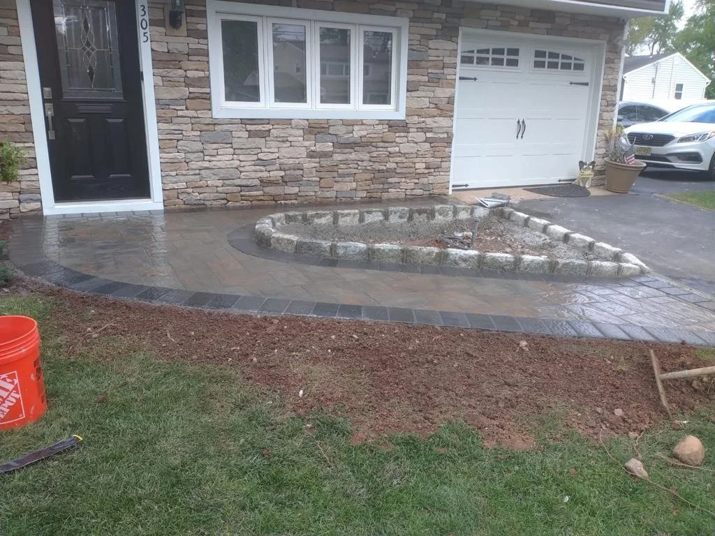 Front yard with freshly paved walkway, partial garden bed with stone border, house with brick facade, white-framed window, black front door, and two cars parked on the driveway.