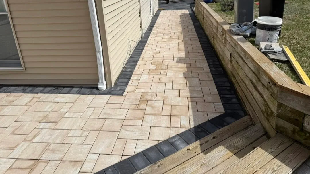 Newly paved beige brick patio with a black border next to a beige siding house; a wooden planter box and construction tools nearby.