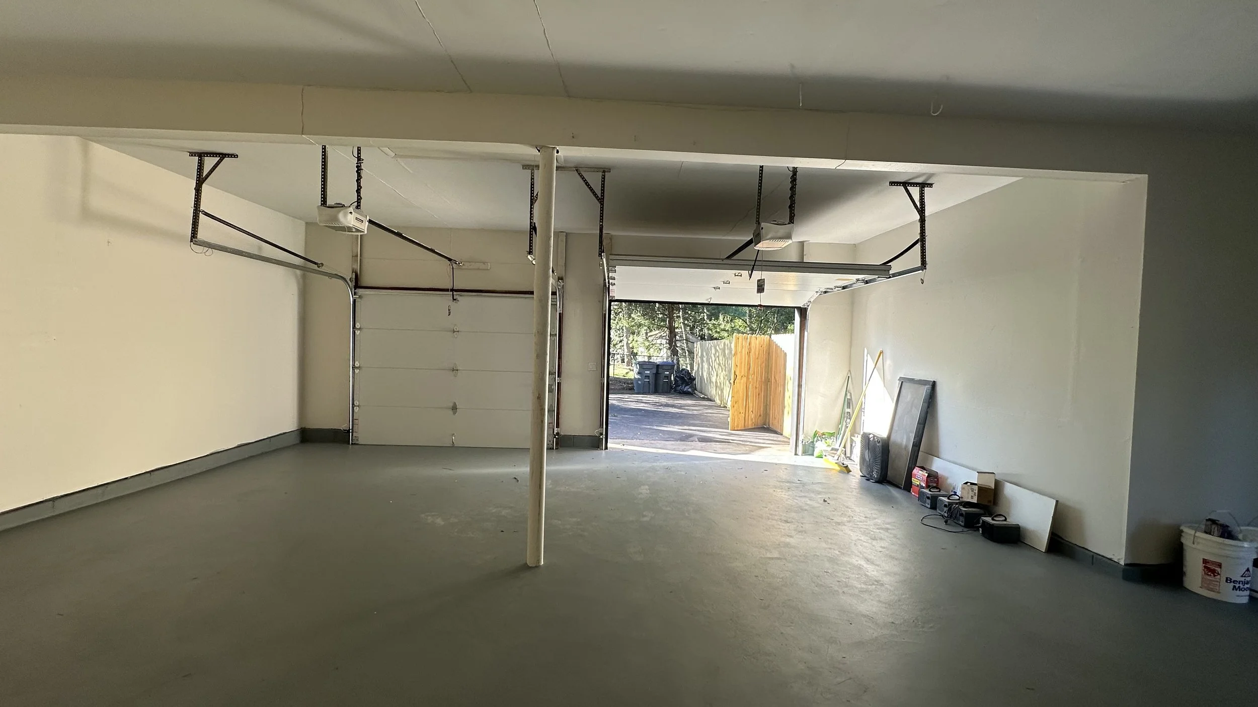 Empty garage with concrete floor, open garage door showing driveway and trees outside, with tools, boxes, and a fan along the right wall.