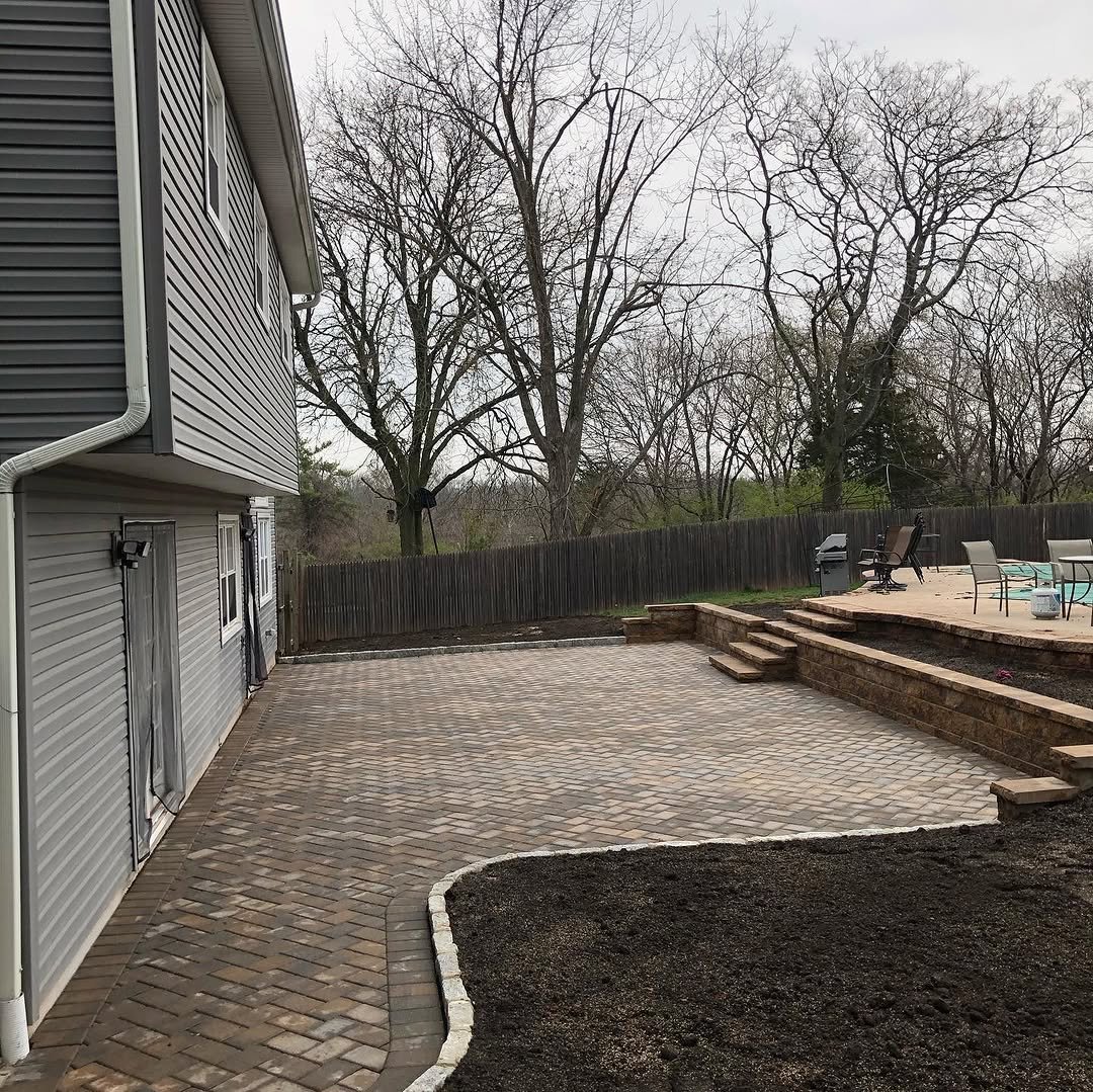 Backyard patio area with brick pavers, a raised section with outdoor furniture and grill, a wooden fence, trees in the background, and the side of a house with vinyl siding.