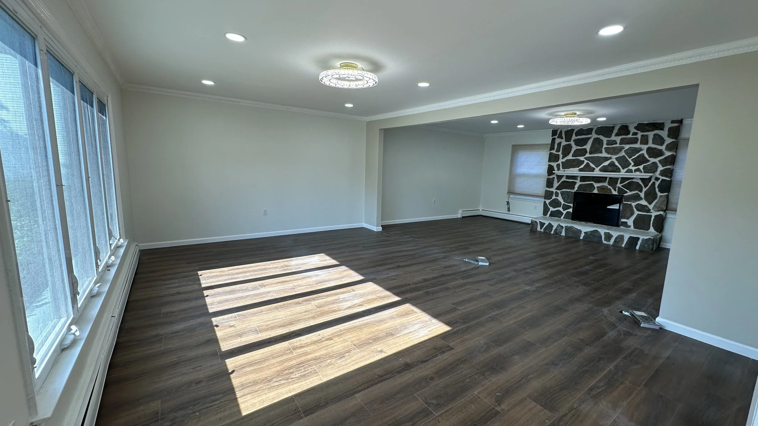 Empty living room with large windows, hardwood floors, a stone fireplace, and ceiling lights.