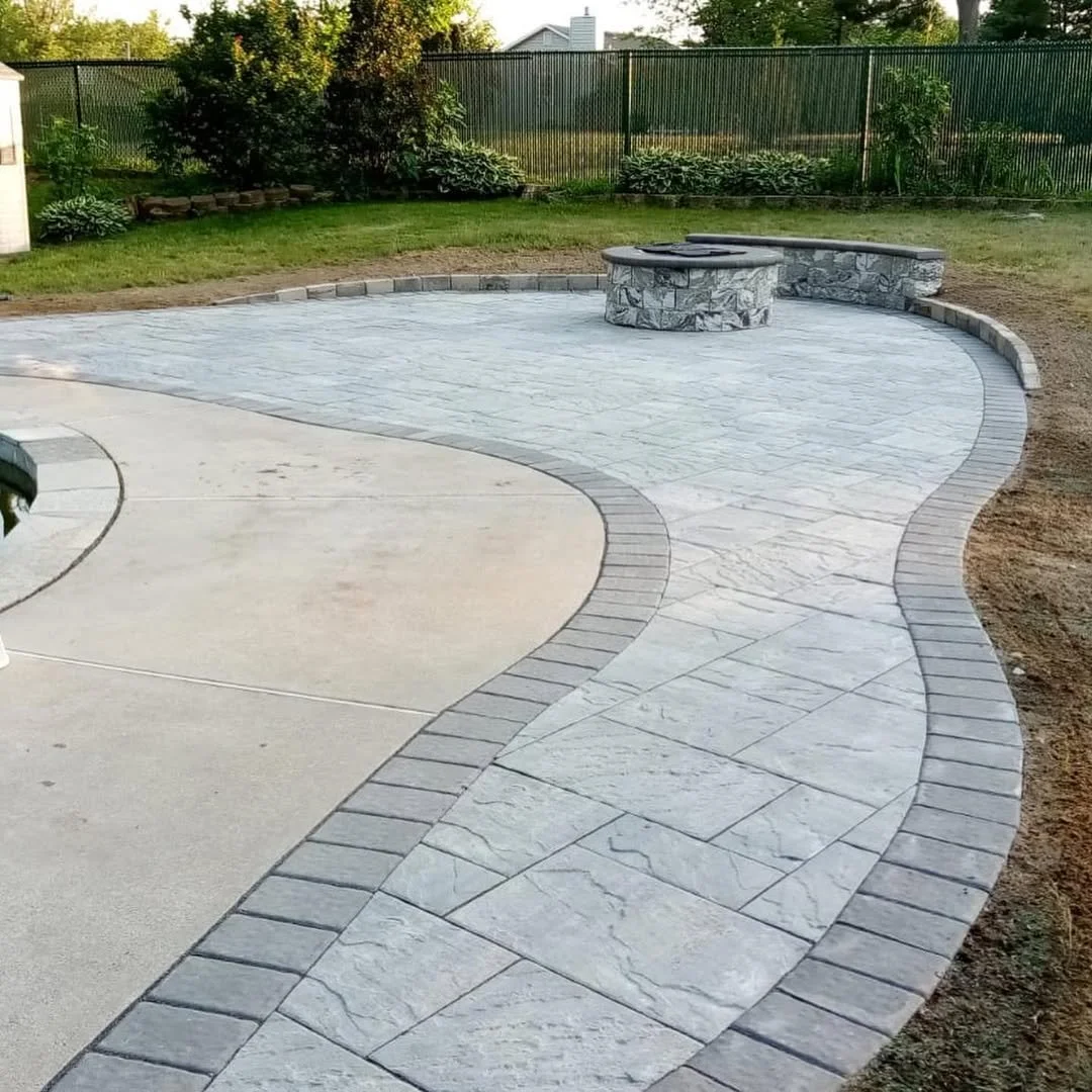 Curved backyard patio with stamped concrete and brick borders, a stone fire pit, surrounded by a lawn and fence.
