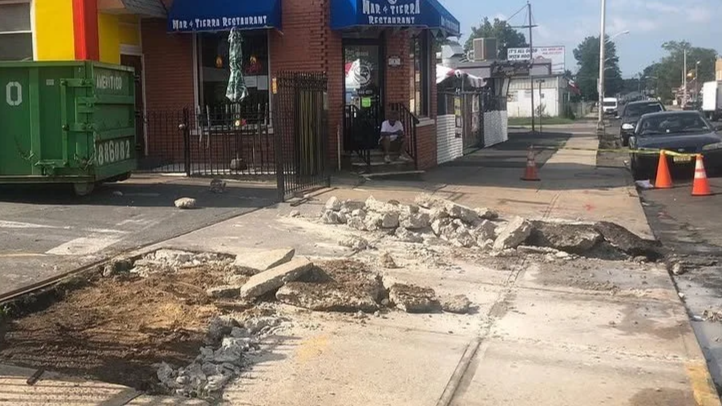 Construction work on a city sidewalk with orange cones, a green dumpster nearby, and a few people outside a brick restaurant with a blue awning.