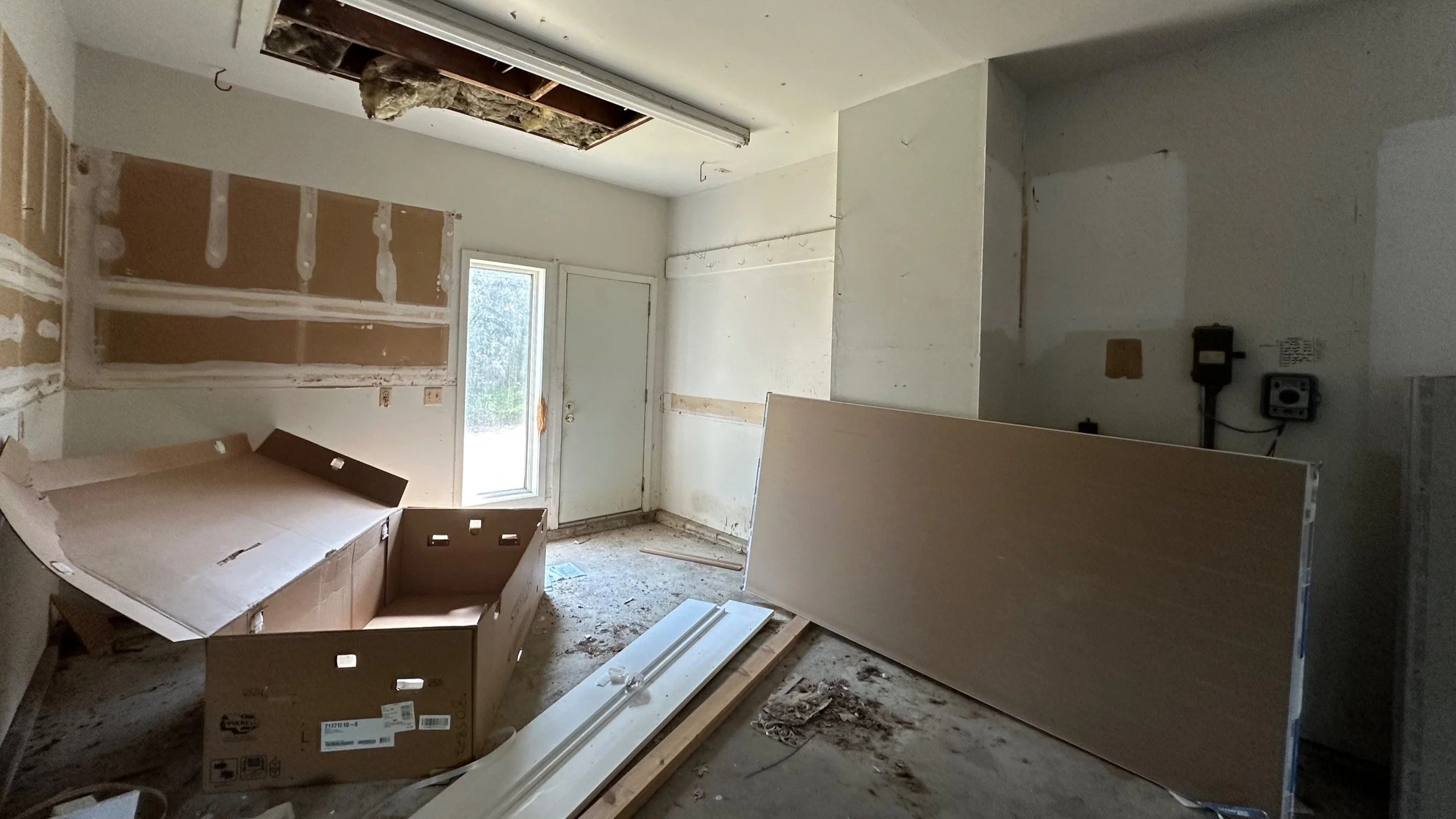 Room under renovation with drywall and ceiling work in progress, scattered cardboard boxes, and construction debris on the floor.