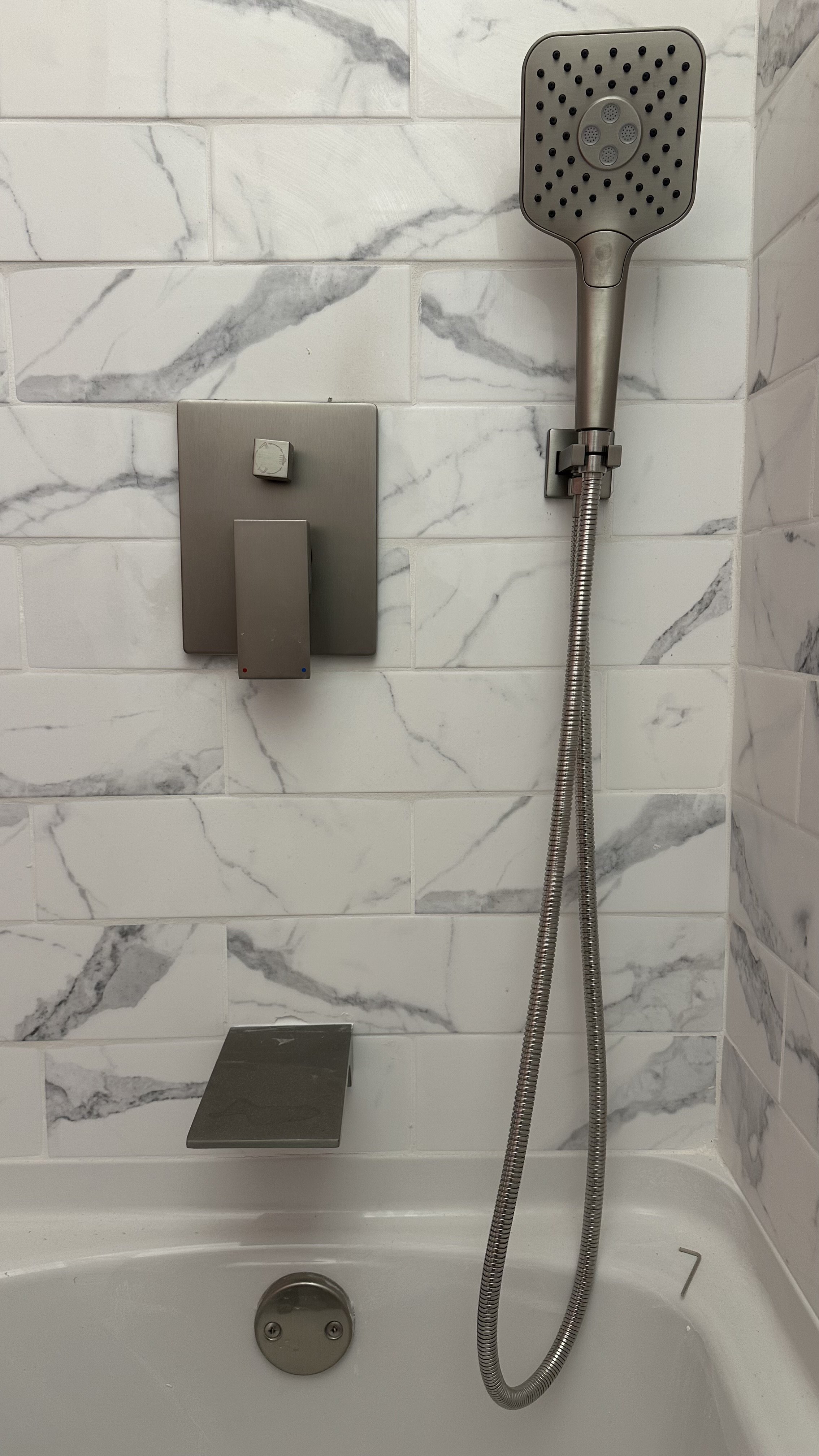 Shower with a grey handheld showerhead, a control panel with a switch, and a small shelf, all mounted on white marble tile wall in a bathroom.