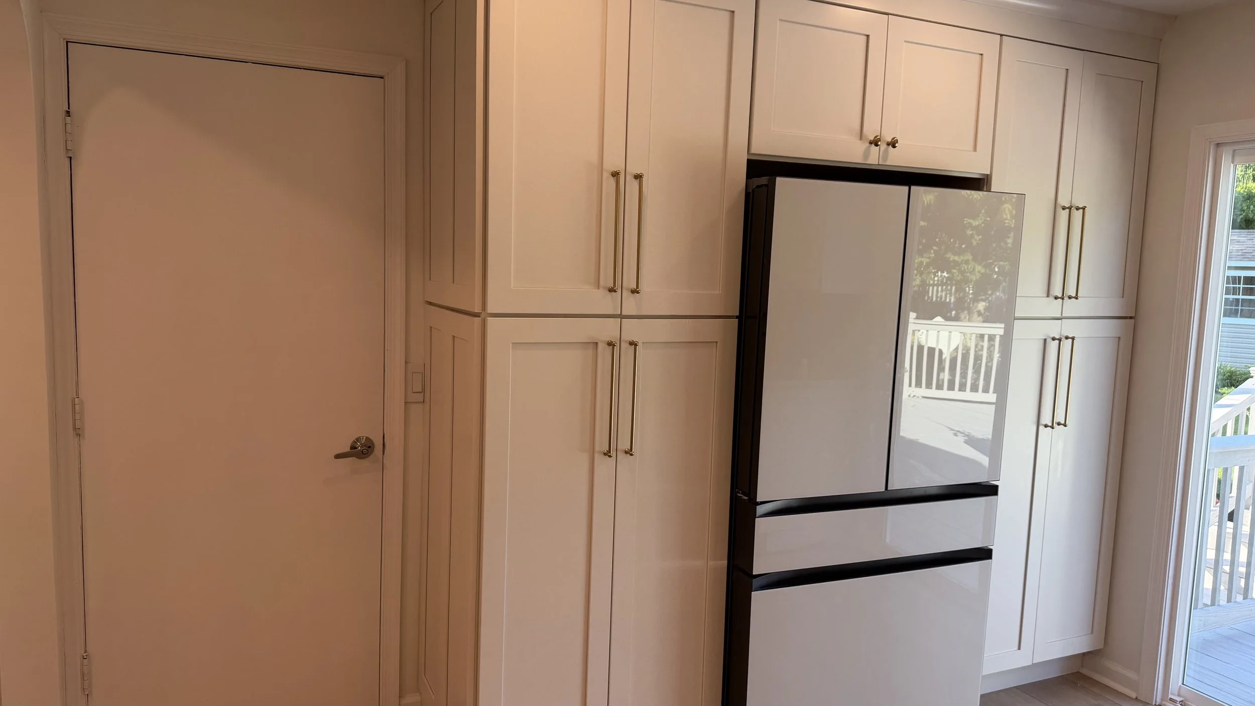 A kitchen with white cabinets and gold handles, a stainless steel refrigerator, and a sliding glass door leading to an outdoor deck with a white railing.
