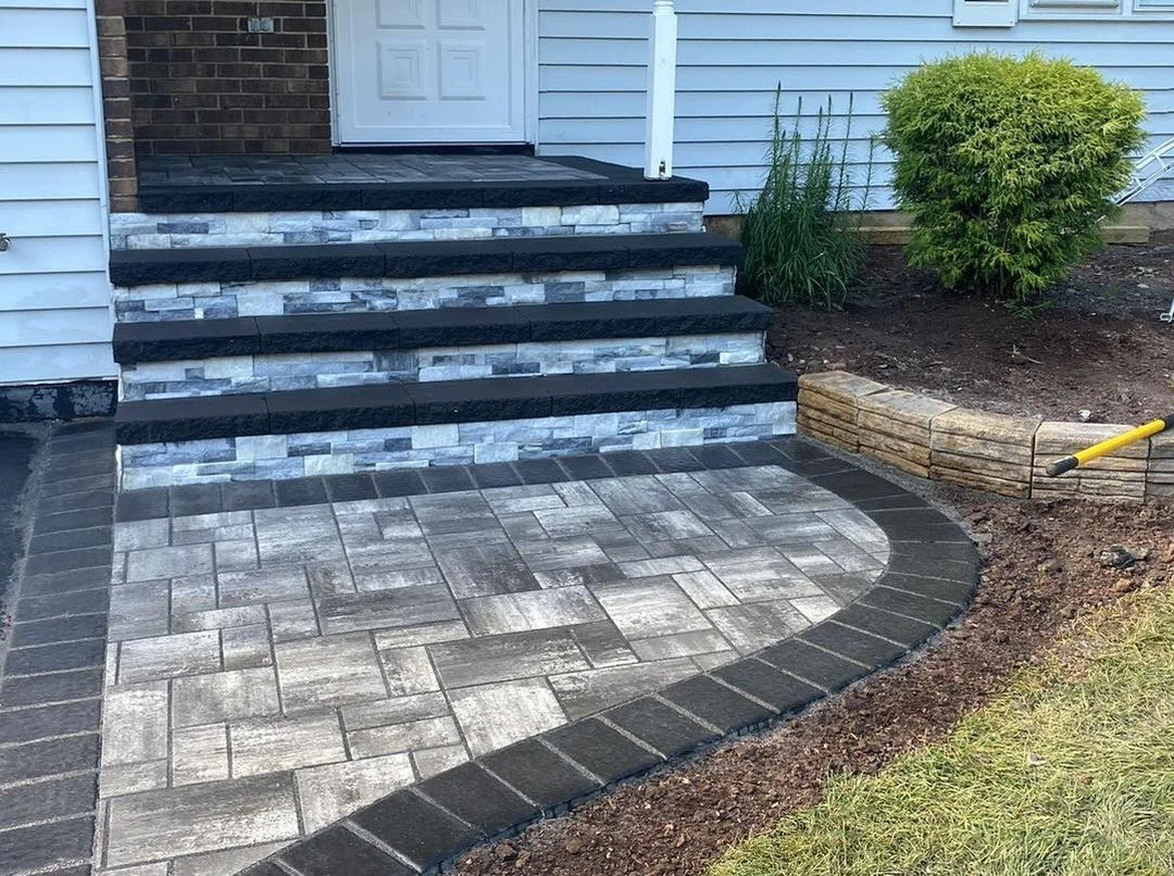Newly paved front porch with stone steps and a multi-colored brick patio, bordered by dark bricks, next to a house with siding, a white door, and landscaped garden beds.
