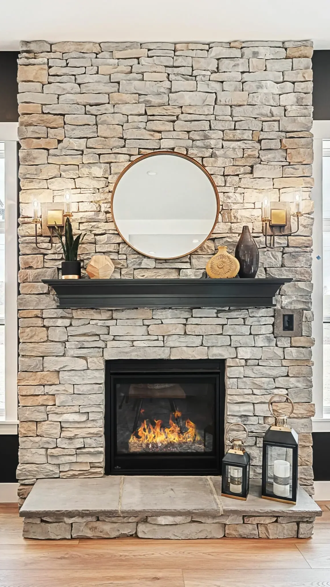 A stone fireplace with a black mantel, decorated with a mirror, vases, and a potted plant. Two candle lanterns sit on the floor in front of the fireplace, which has a fire burning inside.