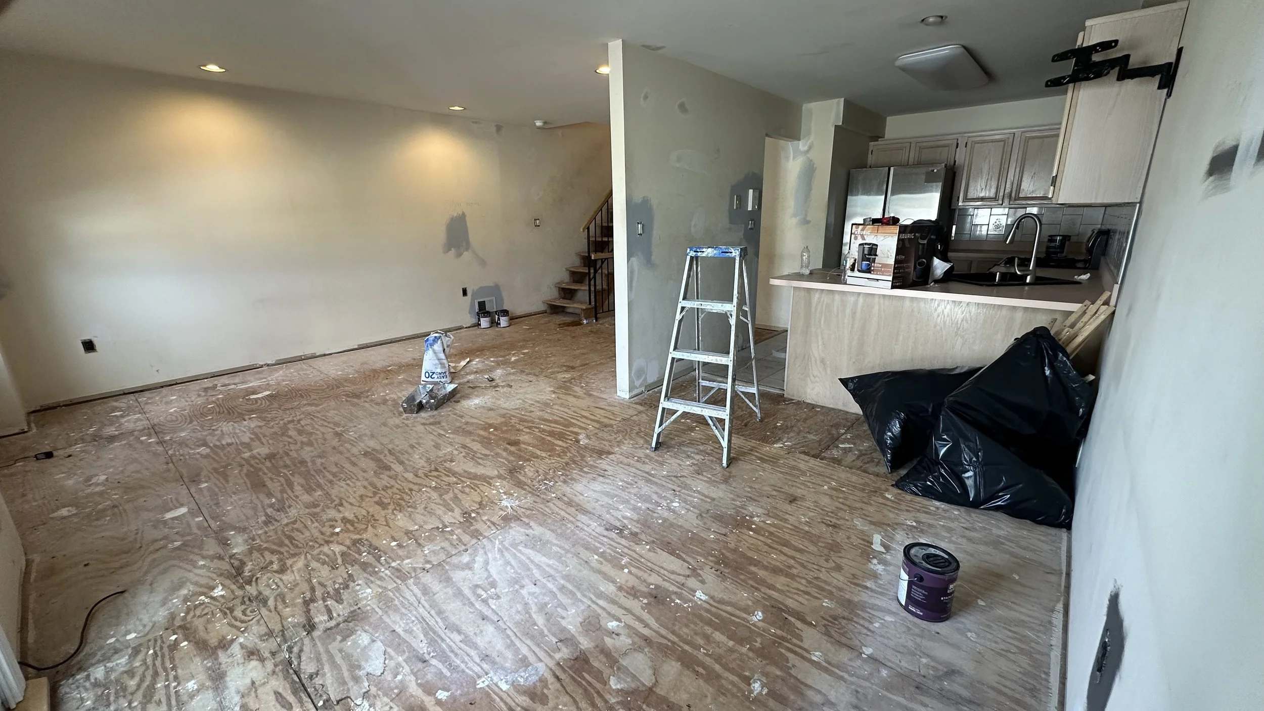 Room undergoing renovation with plywood flooring, a ladder, construction supplies, and partially painted walls.