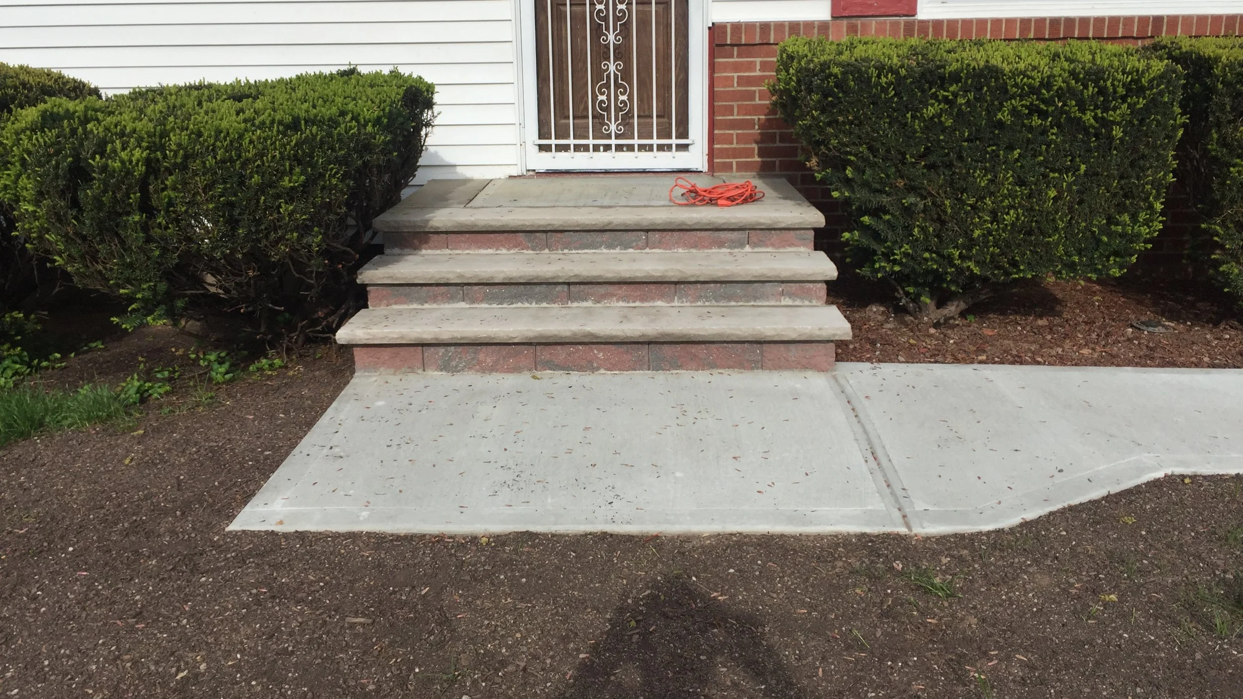 Concrete steps leading up to a red brick and white siding house door, with greenery on either side and a coiled orange extension cord on the top step.