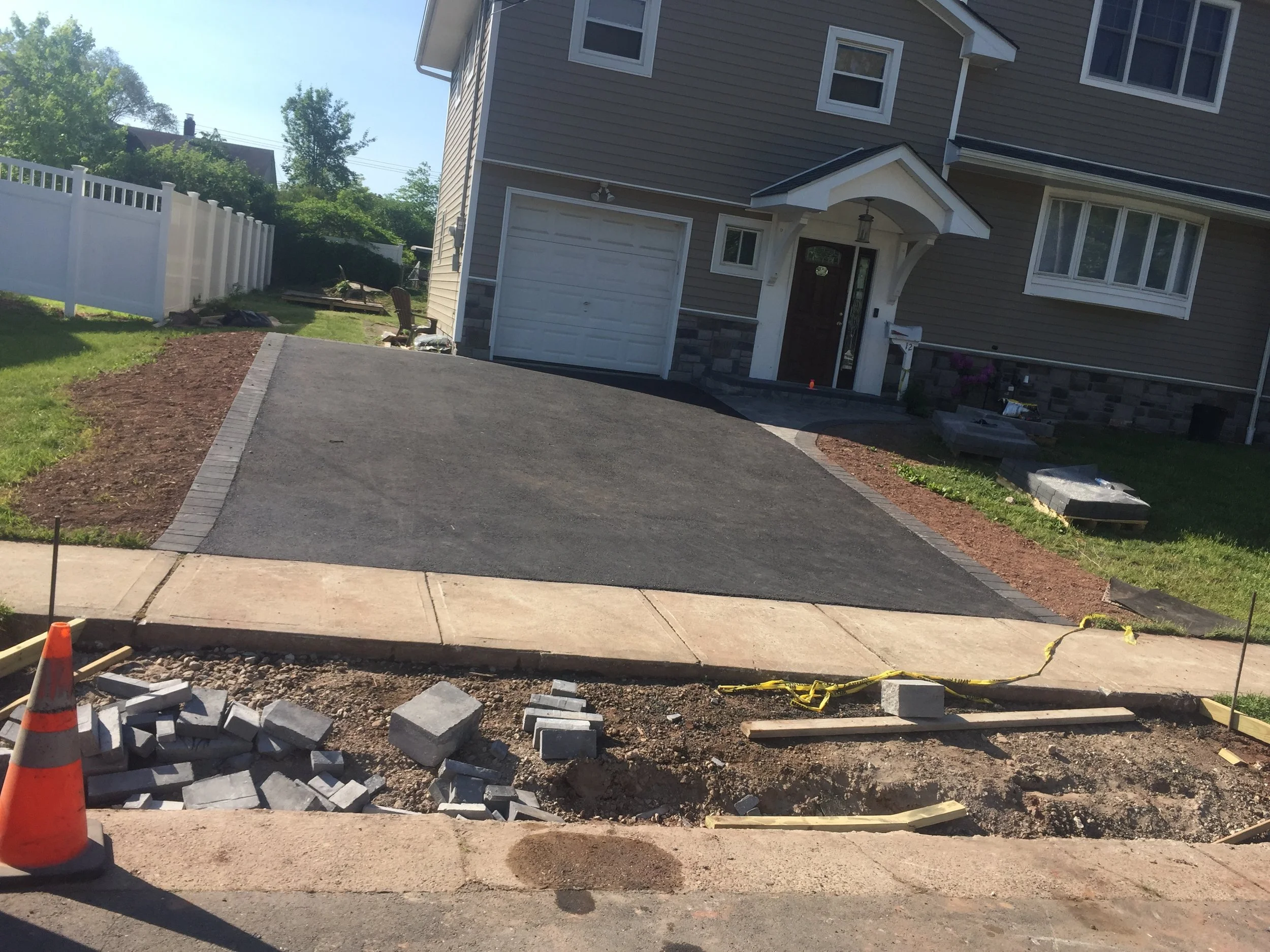 Residential driveway under construction with freshly paved surface, construction cone, and paving materials. House with a garage and front entrance visible in the background.
