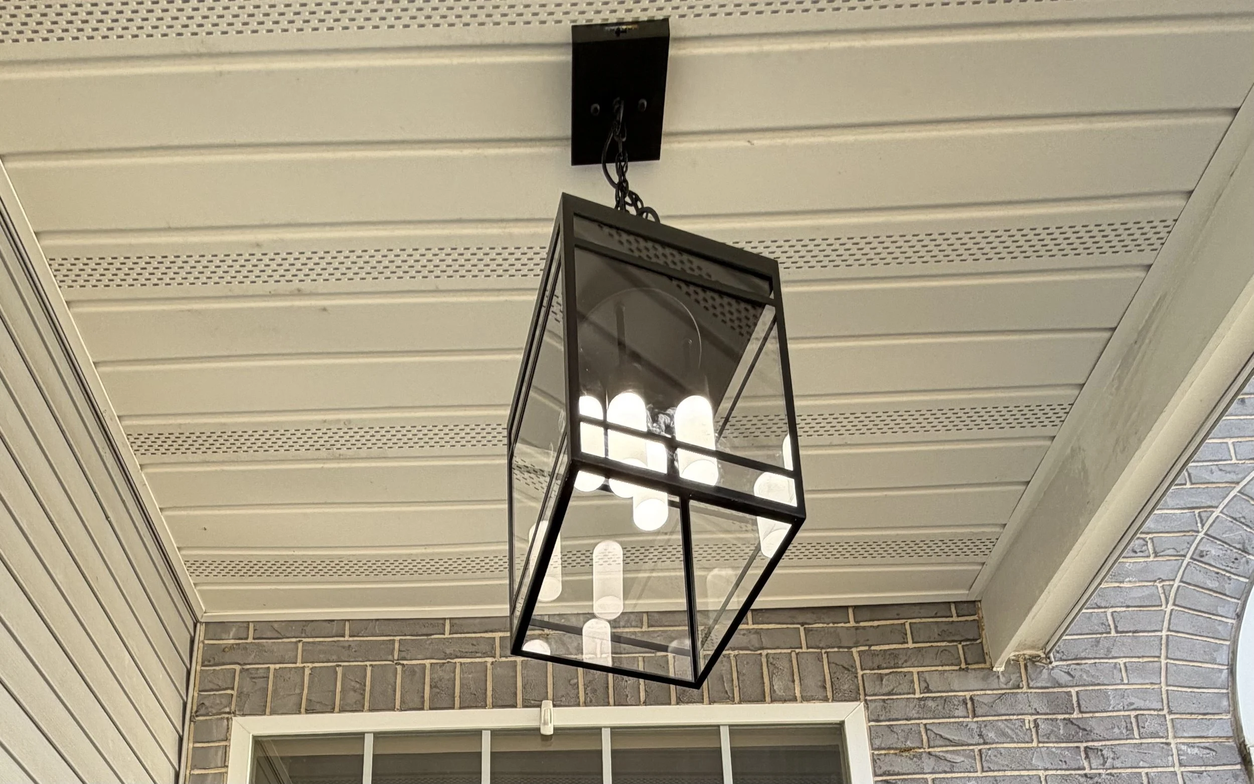 A black metal-framed lantern-style light fixture hanging from a chain, with multiple bulb fixtures inside, mounted on a ceiling with white vinyl paneling and a brick wall below.