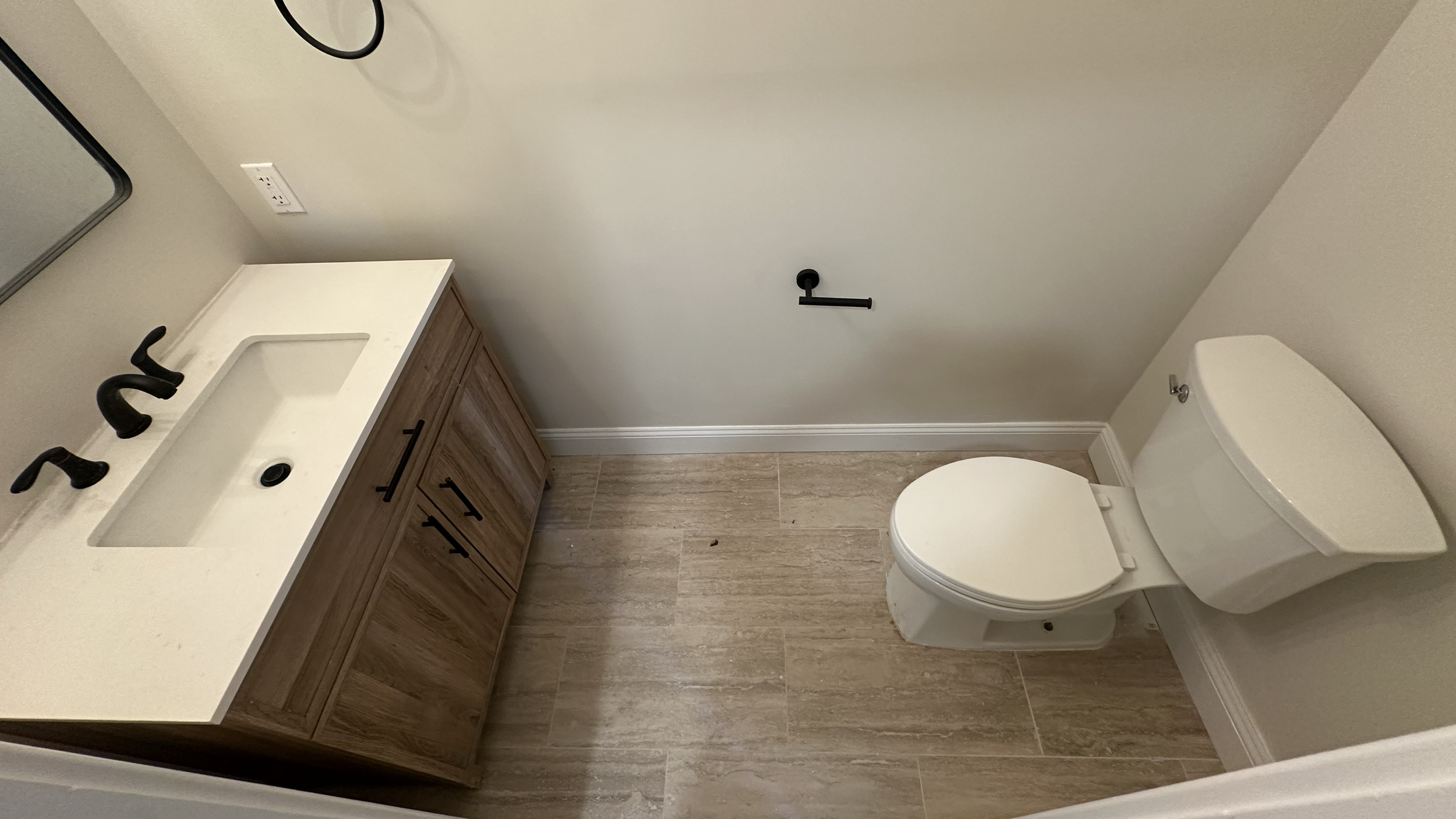 A small bathroom with a wooden vanity with a white countertop and a built-in sink, a black faucet, a mirror, a white toilet, a black toilet paper holder, and tiled floor and walls.