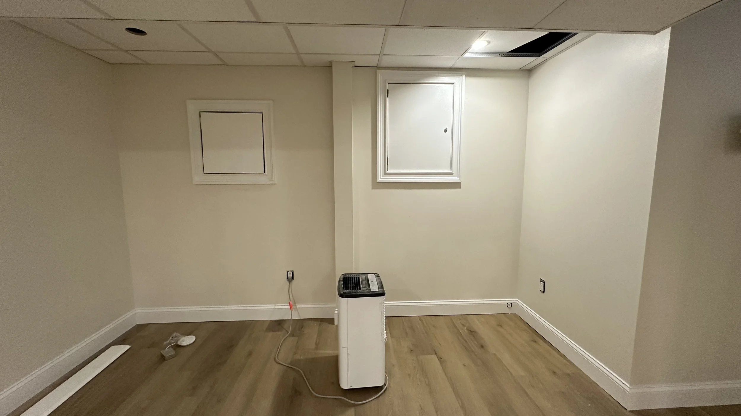 Empty room with beige walls, hardwood floor, small window, and portable air conditioner in the center.