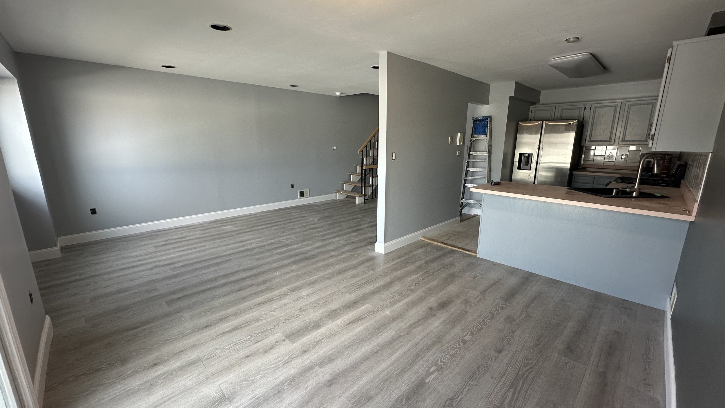 Empty living room and kitchen with light flooring, gray walls, and stainless steel appliances.