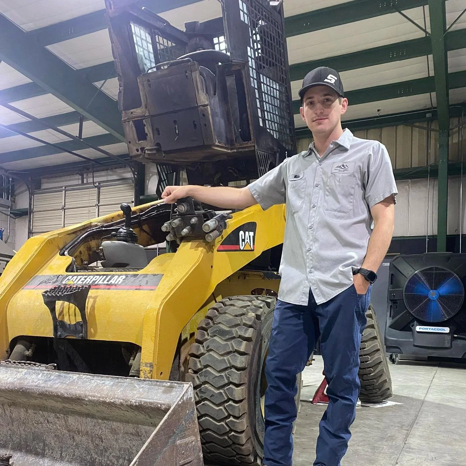 Young man in work attire standing next to a yellow Caterpillar forklift inside a warehouse.