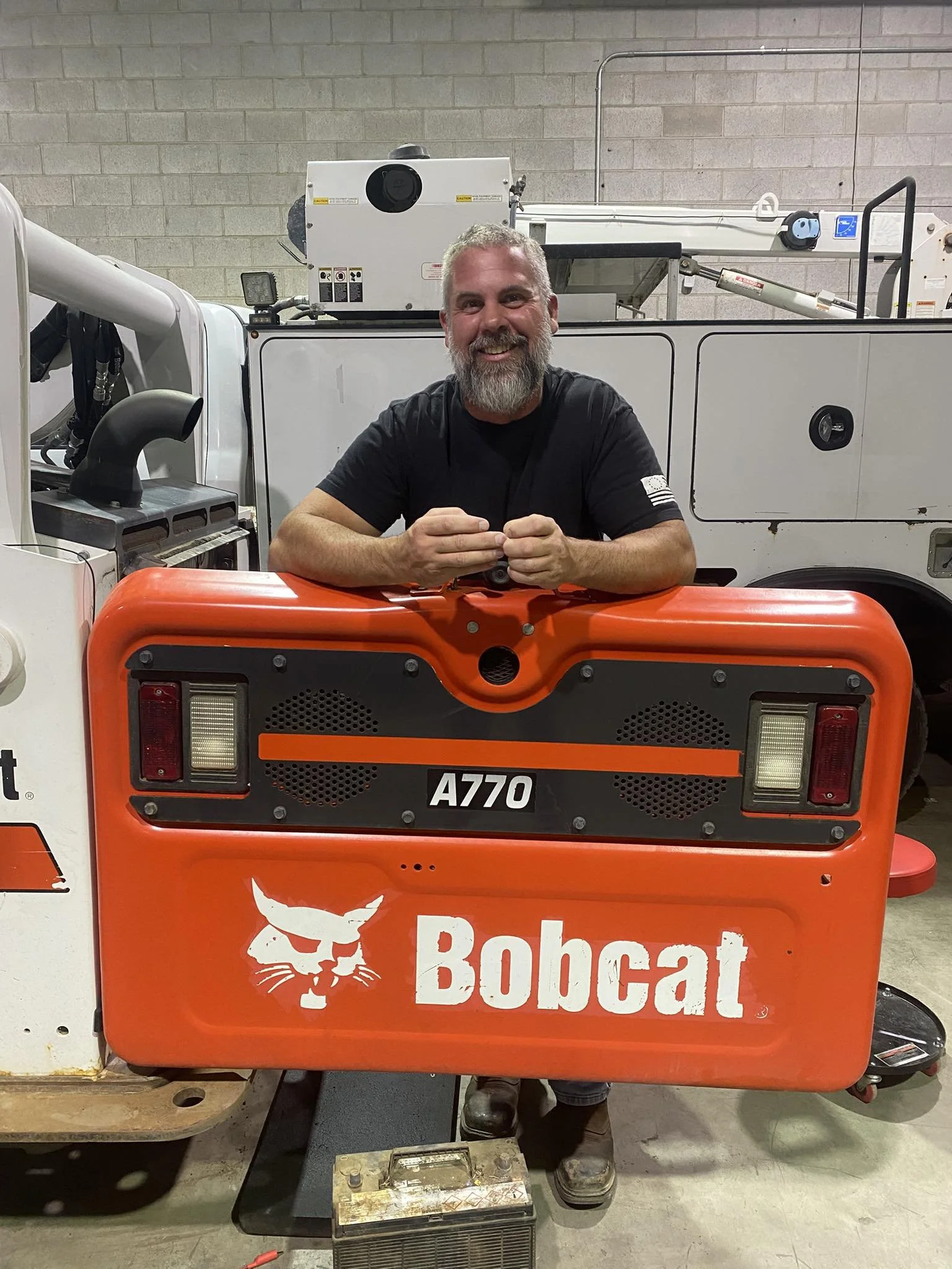 A smiling man with a beard is sitting behind a bright orange Bobcat skid-steer loader inside a warehouse or workshop with concrete block walls.