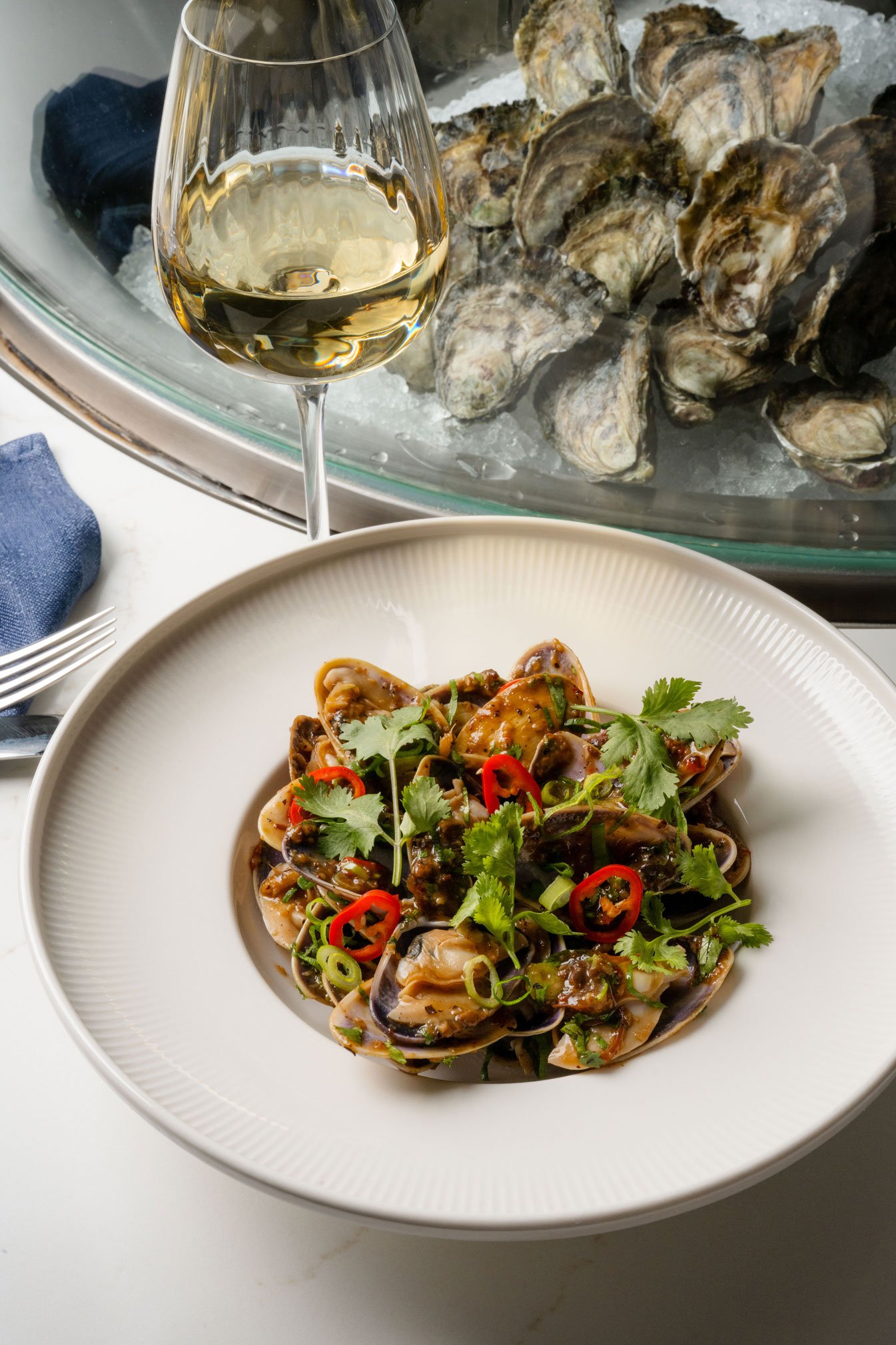 A plate of seafood pipis garnished with cilantro and sliced red chili peppers, with a glass of white wine and a tray of oysters in the background.