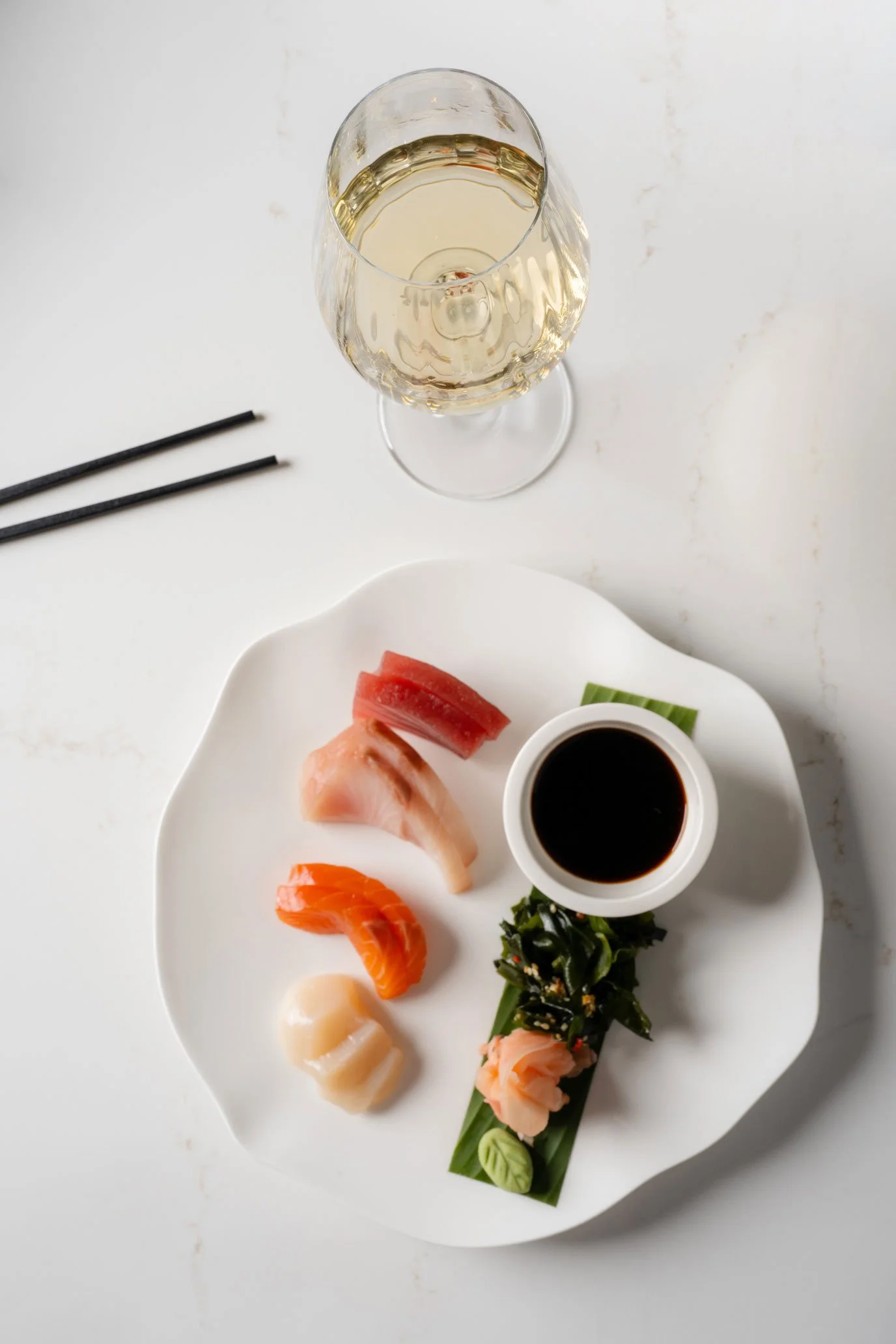 A white plate with assorted sashimi, a small cup of soy sauce, a glass of white wine, and black chopsticks on a white surface.