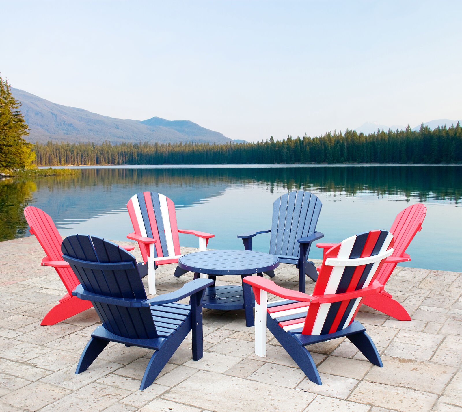 Six Adirondack chairs in red, white, and blue arranged around a matching round table on a stone patio by a calm lake, with mountains and a forest in the background on a clear day.