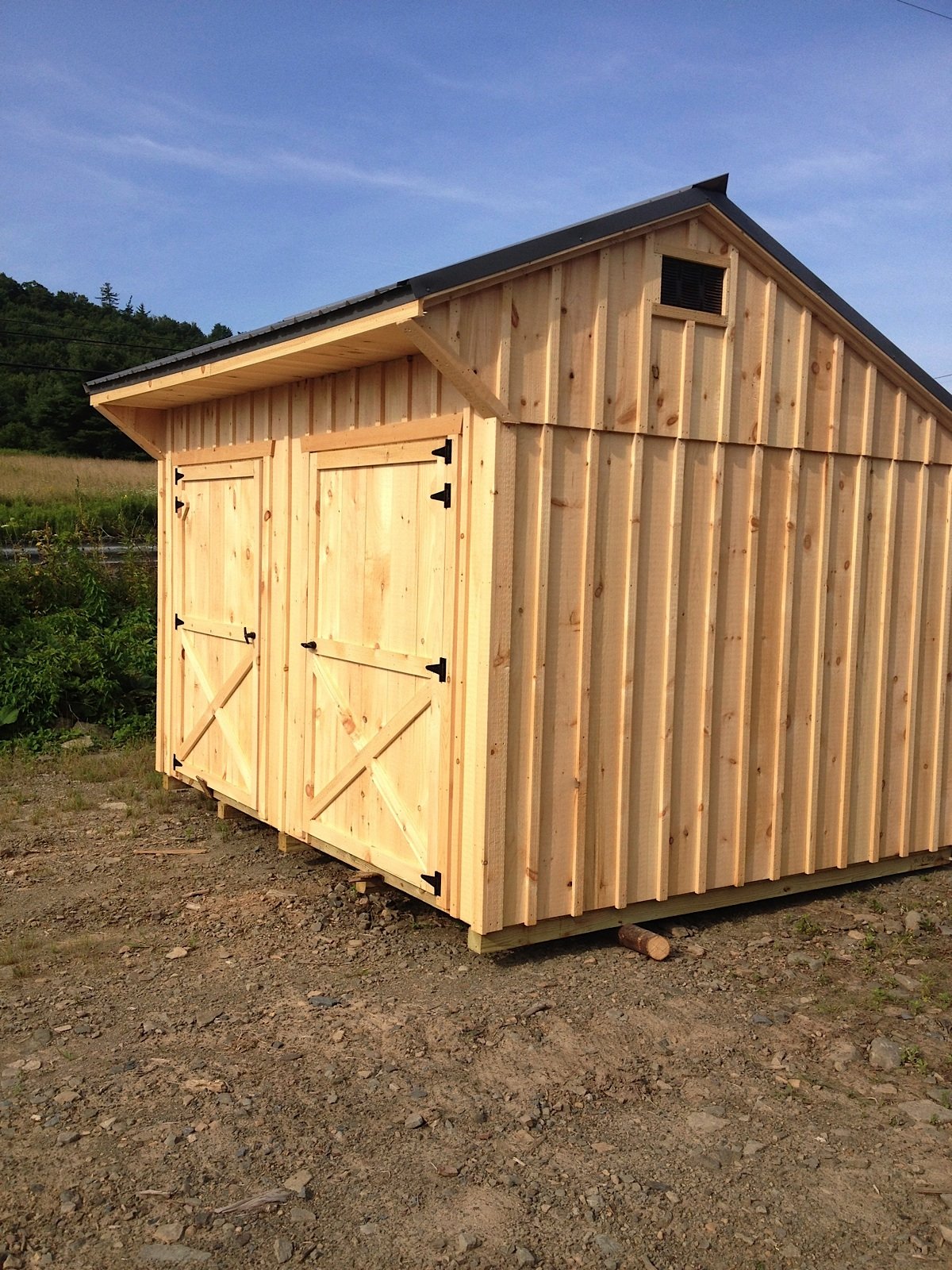 Unfinished wooden shed with double doors, located outdoors on uneven ground with grass and trees in the background under a blue sky.