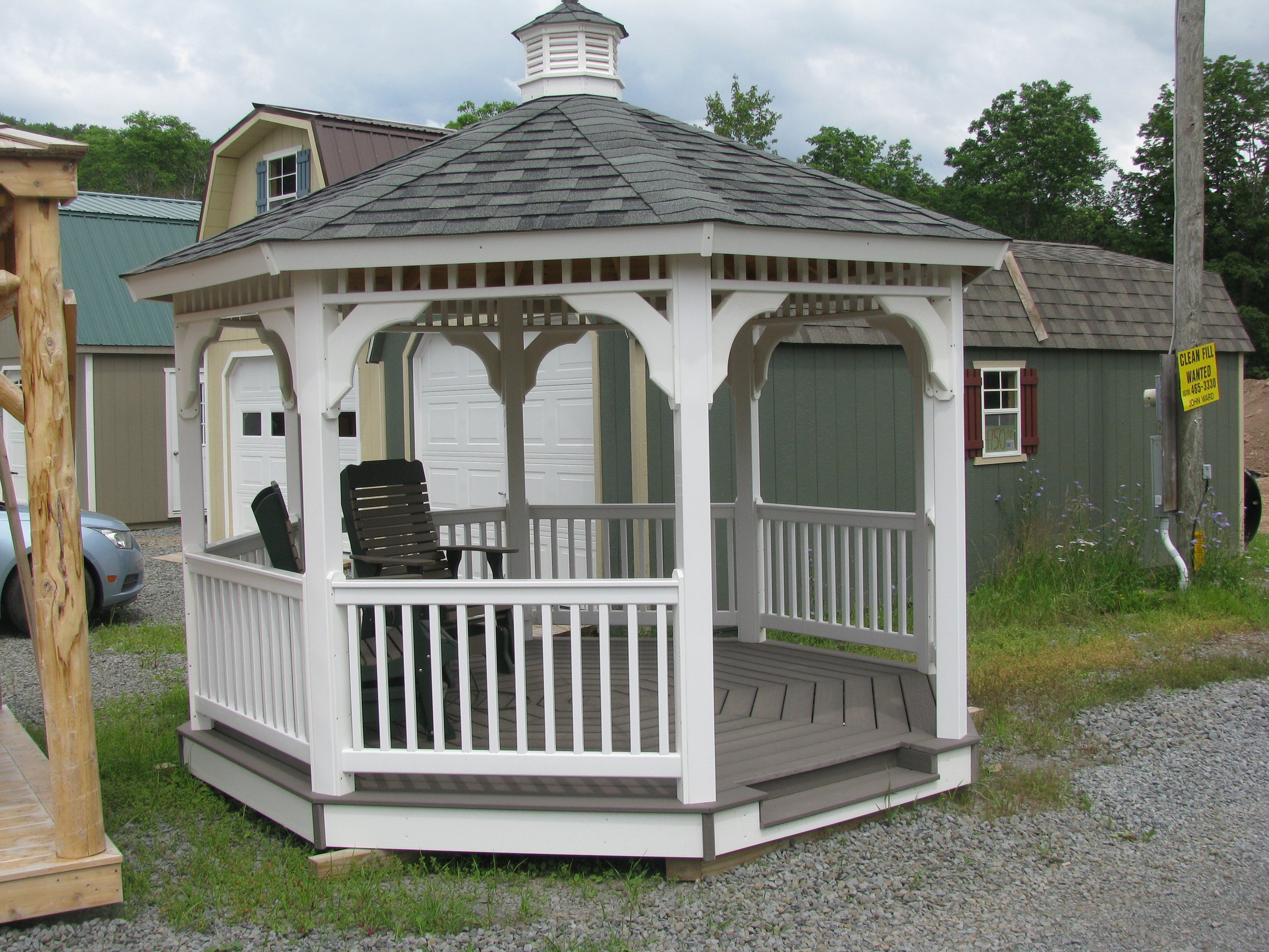A white wooden gazebo with a shingled roof and built-in chairs, outdoors on a gravel surface, with small buildings and trees in the background.