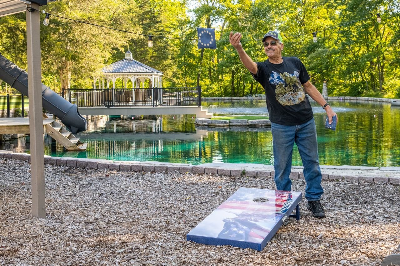 An older man playing cornhole outdoors by a pond, with a European Union flag on the bean bag, wearing sunglasses, a black t-shirt, jeans, and a cap, in a park with trees and a gazebo.