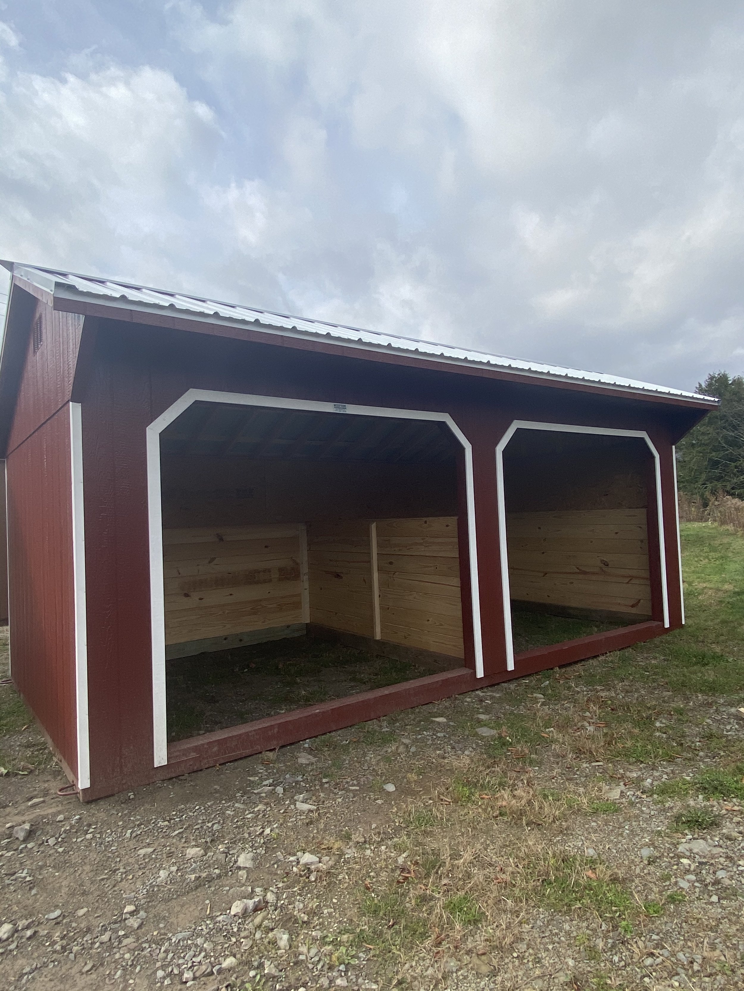 A red horse barn with white trim, open front, and wooden interior walls, on a gravel and grass ground under a cloudy sky.