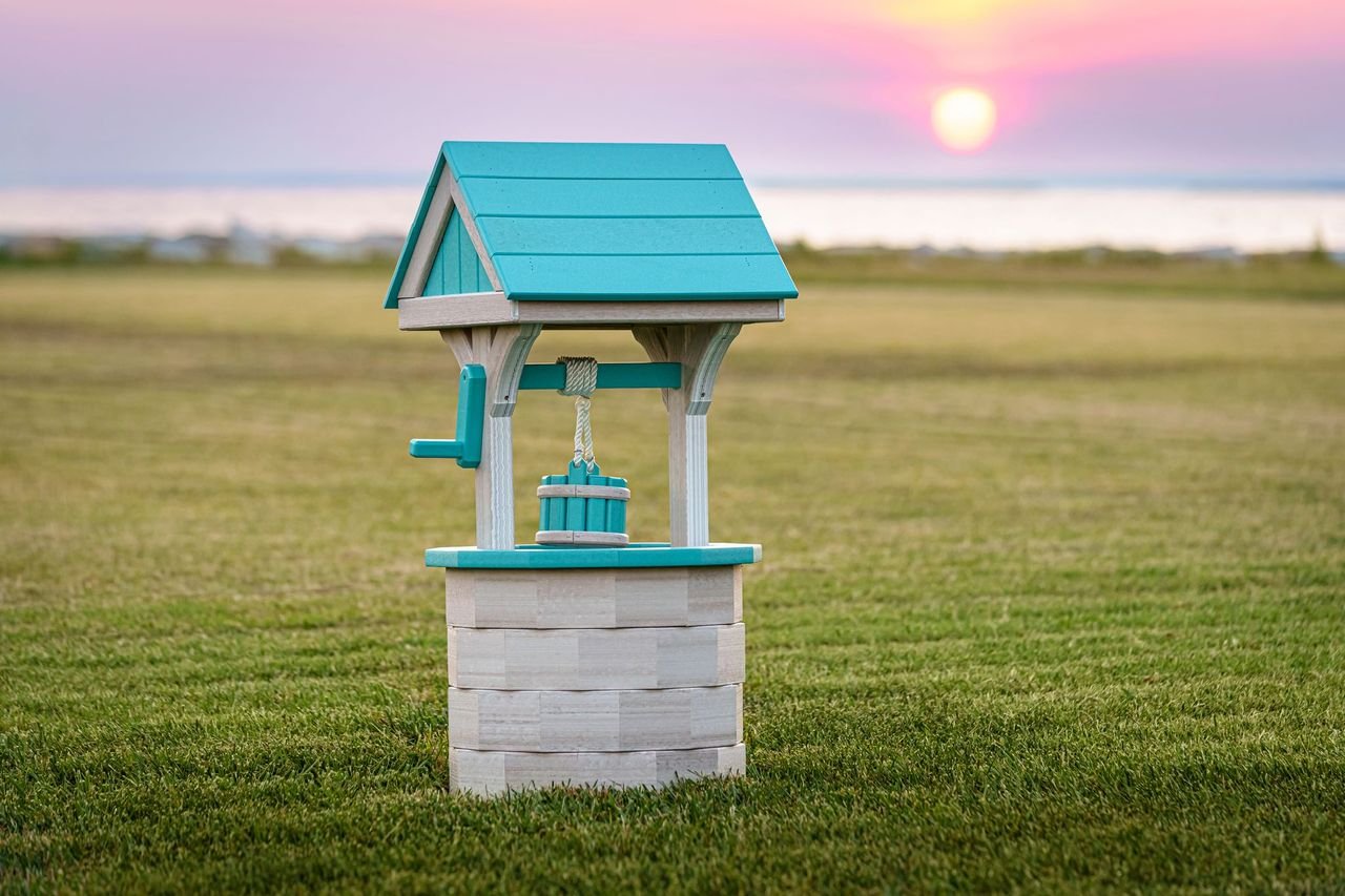 A small decorative well with a blue roof and a bucket hanging on a rope, set on a grassy field during sunset.