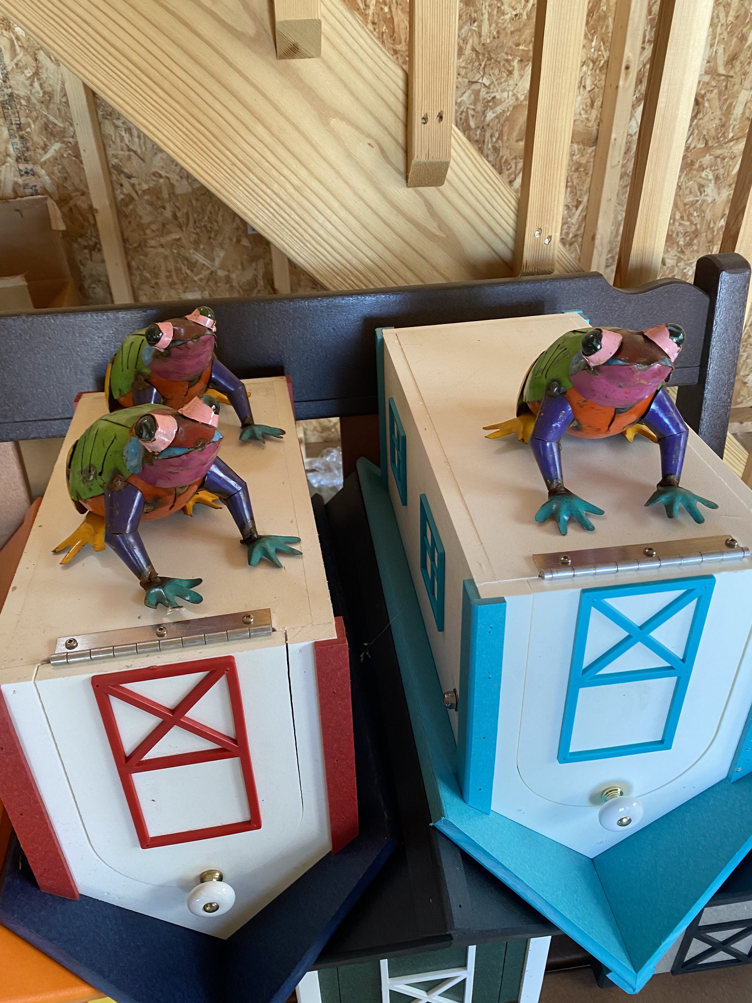 Decorative frog figurines on top of two small wooden barn-style house decorations, with a background of a wooden staircase and unfinished wall. The frogs are colorful, sitting on the roofs of the miniature houses.