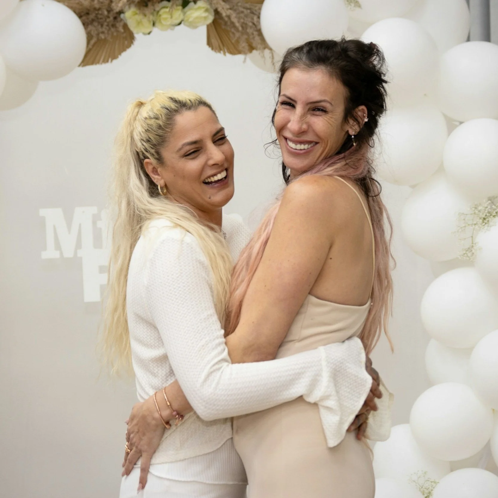 Two women smiling and hugging each other in front of a white balloon backdrop.