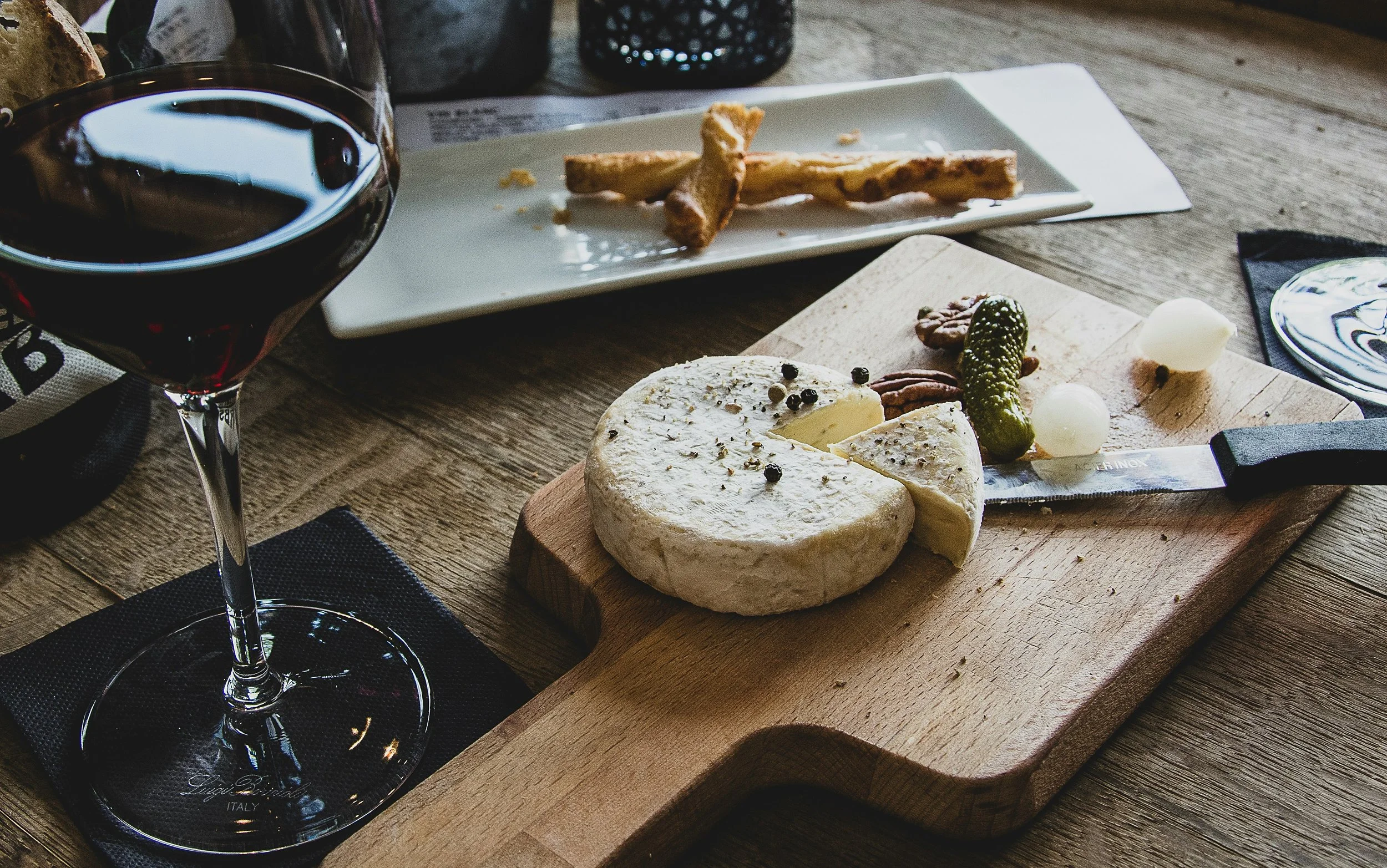 A glass of red wine, a cheese wheel with a slice cut, a knife, and a platter of breadsticks on a wooden table.
