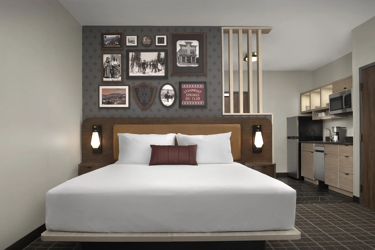 Hotel room with two beds, a glass-enclosed workspace, a sitting area with a tray table, and a large window with curtains.