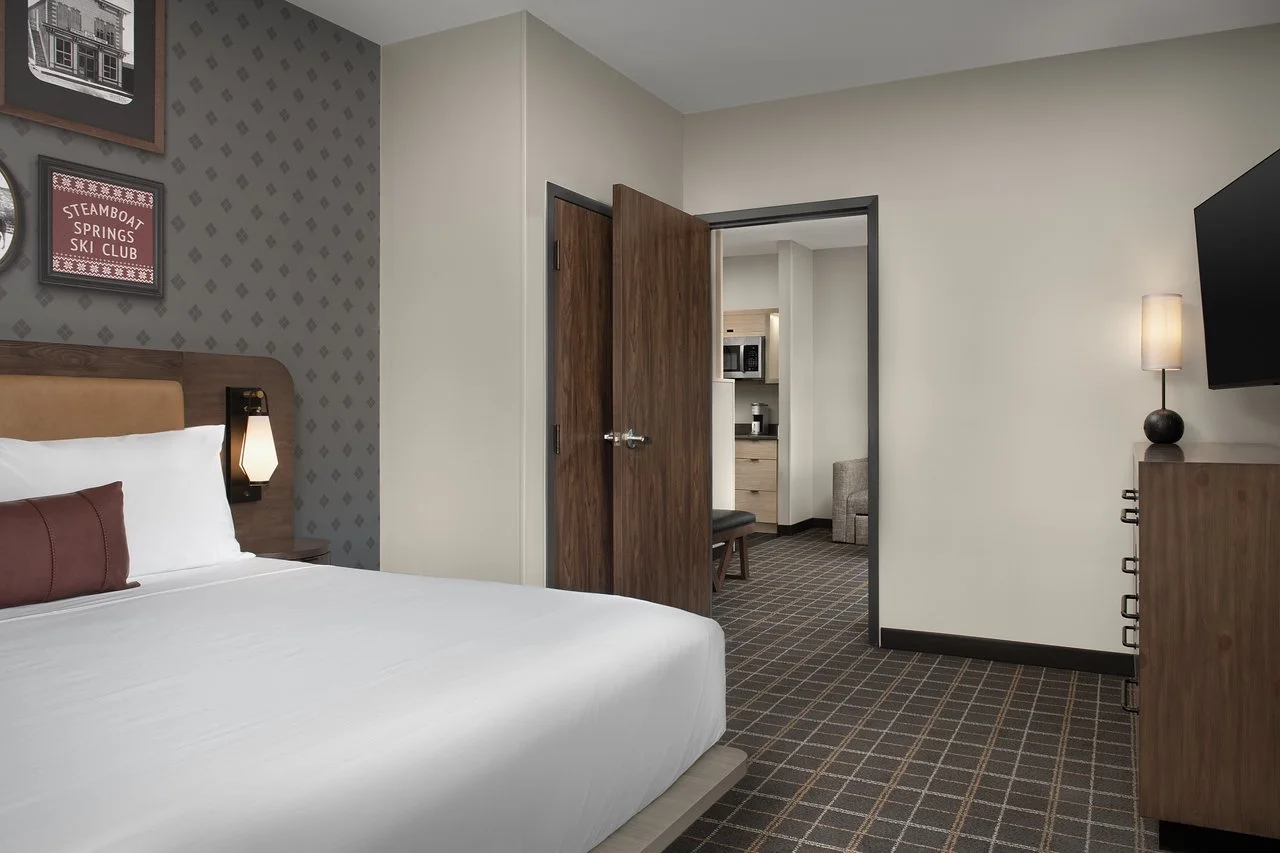Hotel room with a bed, nightstand, lamp, wooden slats, kitchen area, and modern decor.